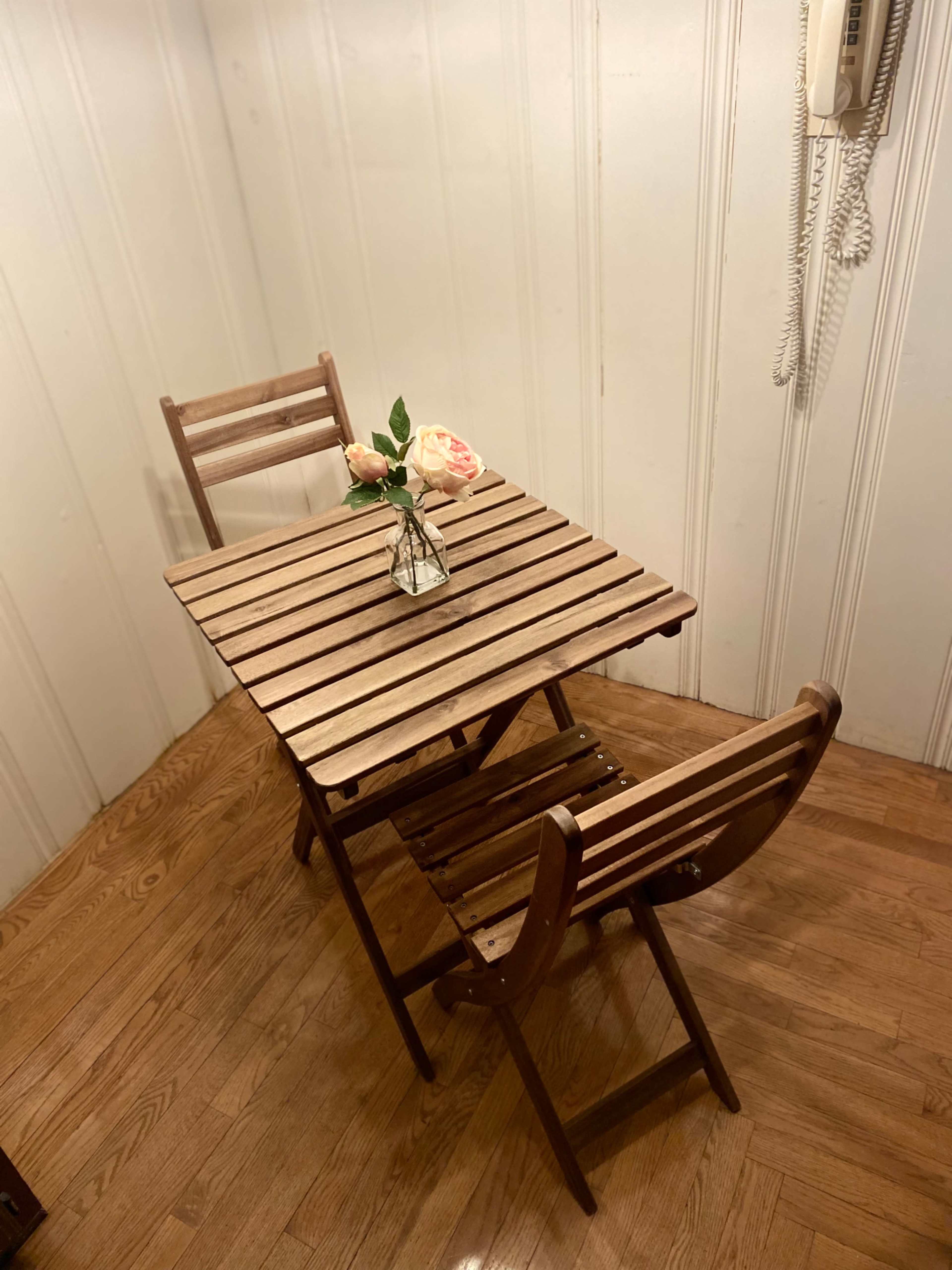 A small wooden table with two folding chairs sits in a corner, featuring a vase of roses on the table.