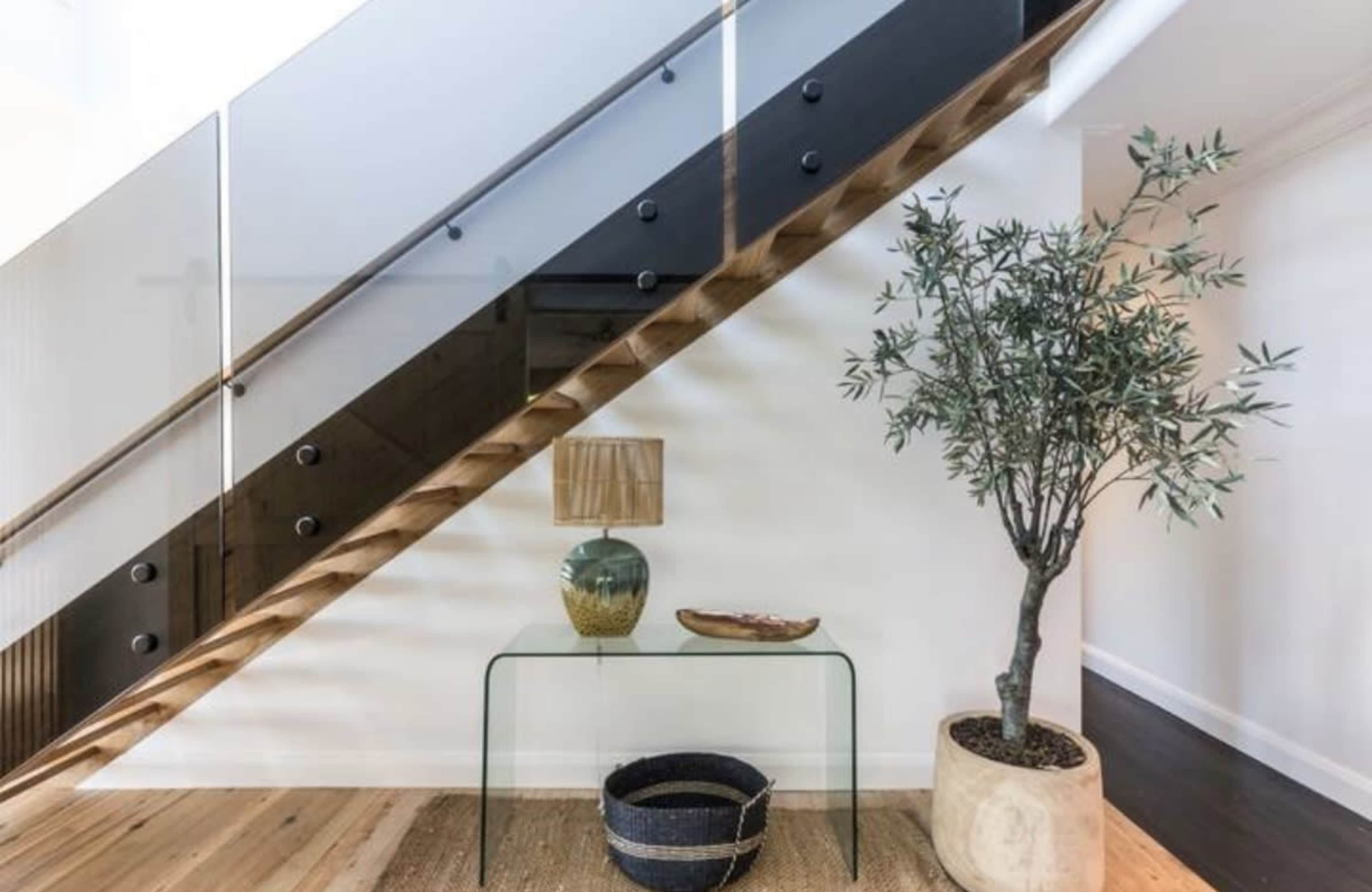 A modern staircase with wooden steps and glass railing features a decorative table with a lamp and a potted plant at its base.