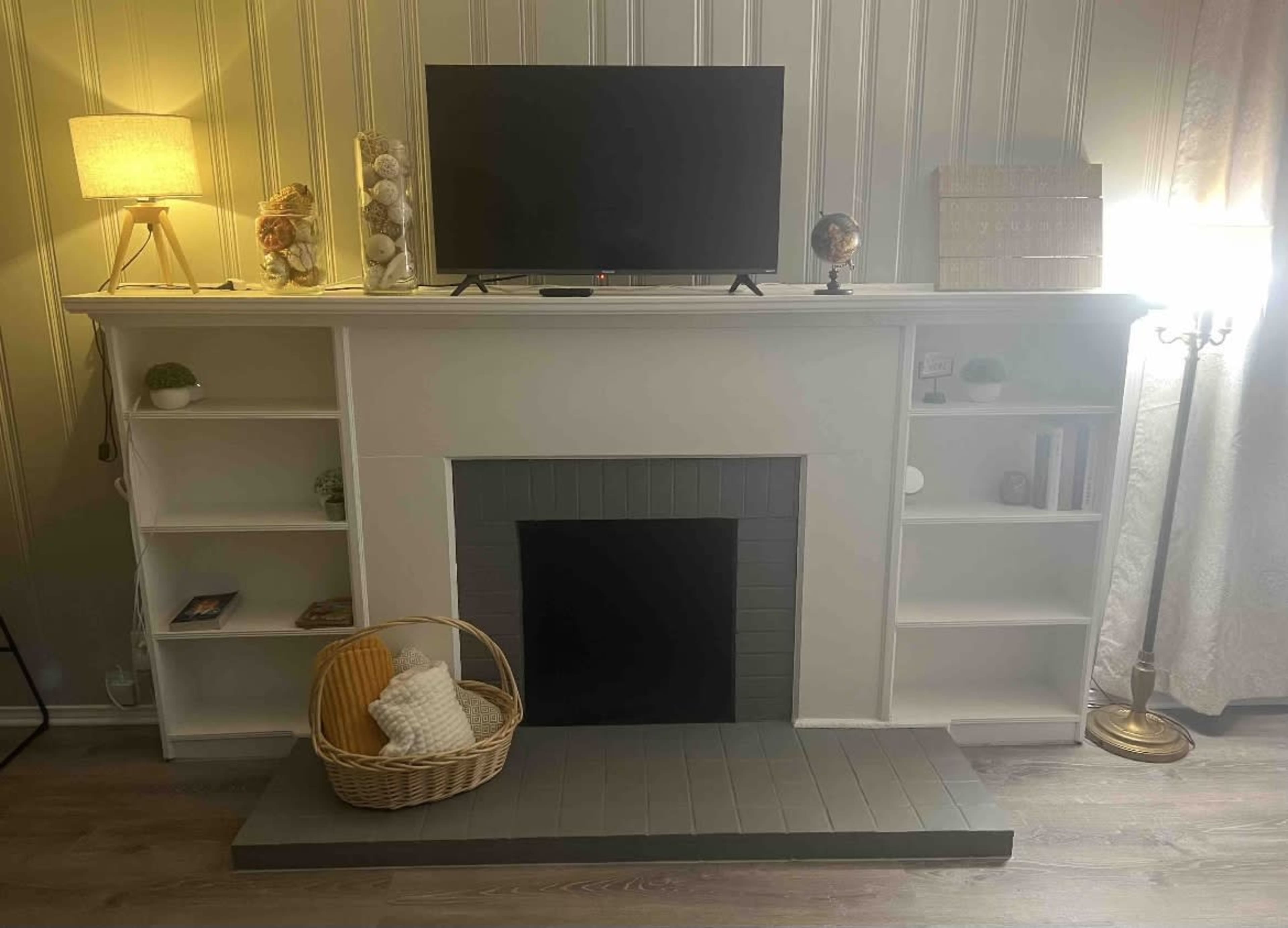 A living room setup featuring a white mantel with a television on top, flanked by shelves, and a basket on the floor beside a fireplace.