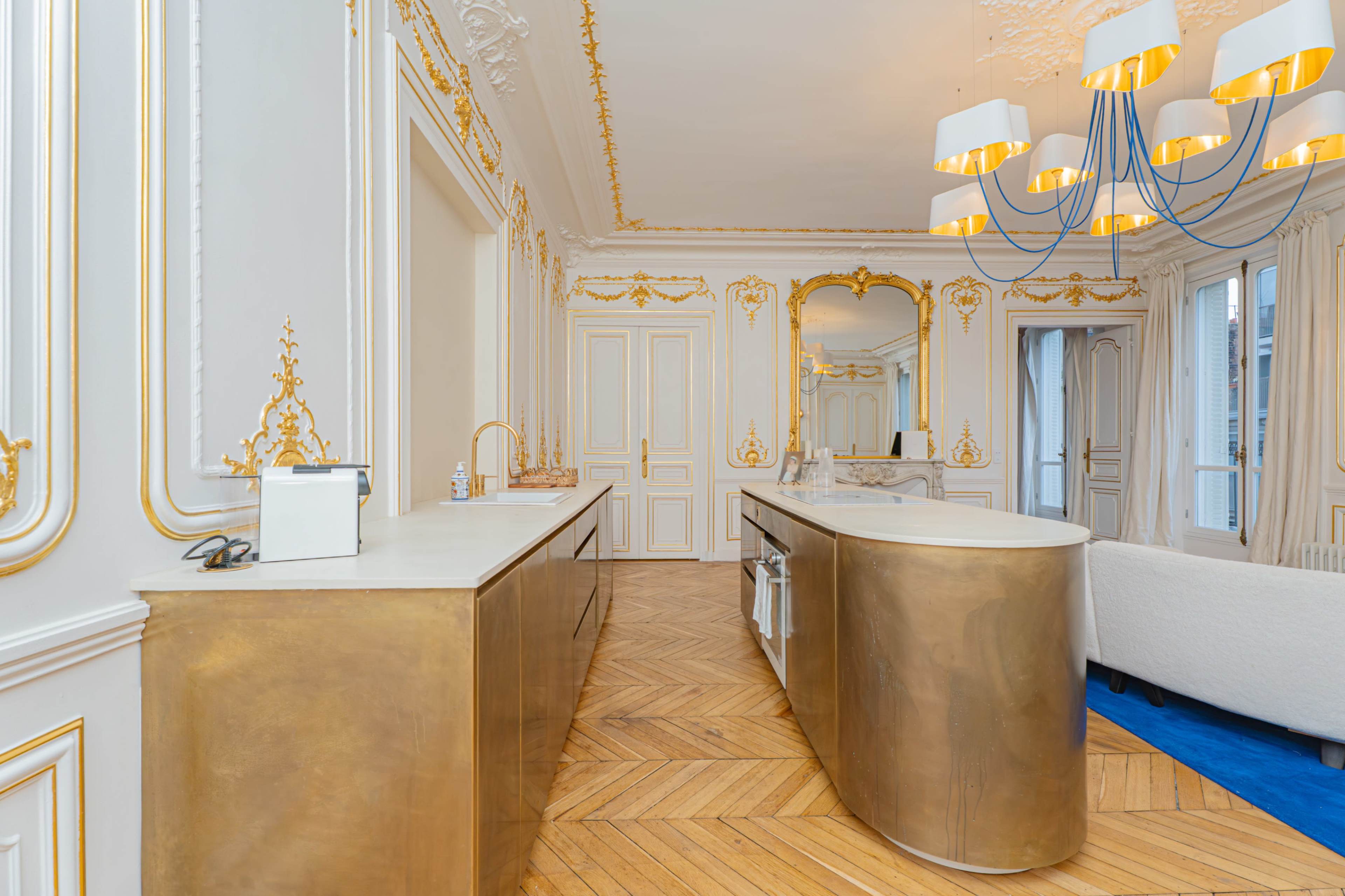The image shows a luxurious kitchen with a gold-accented island, ornate moldings, and elegant lighting fixtures.