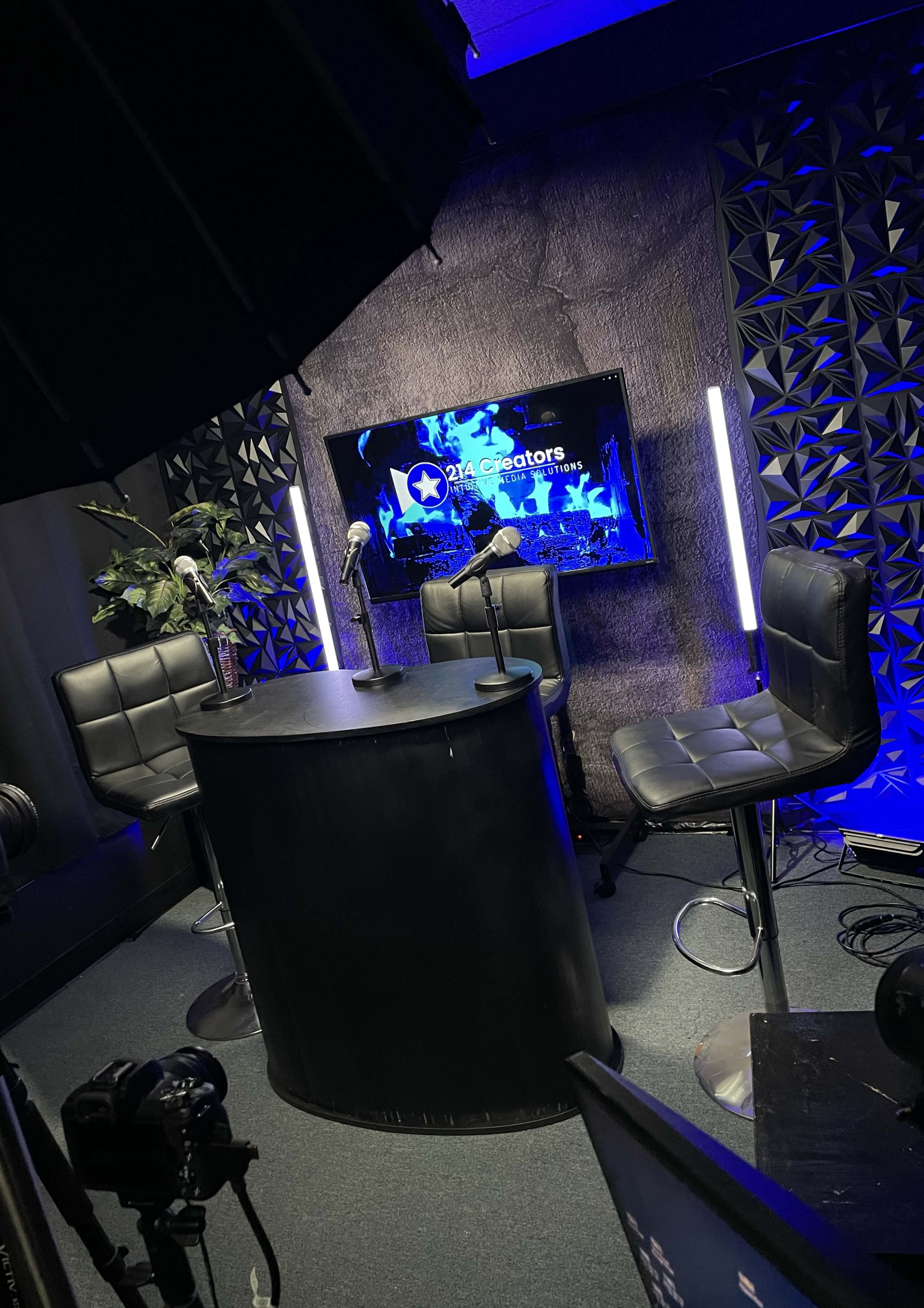 A recording studio setup with a round table, three bar stools, a backdrop featuring a screen with creative graphics, and lighting elements.