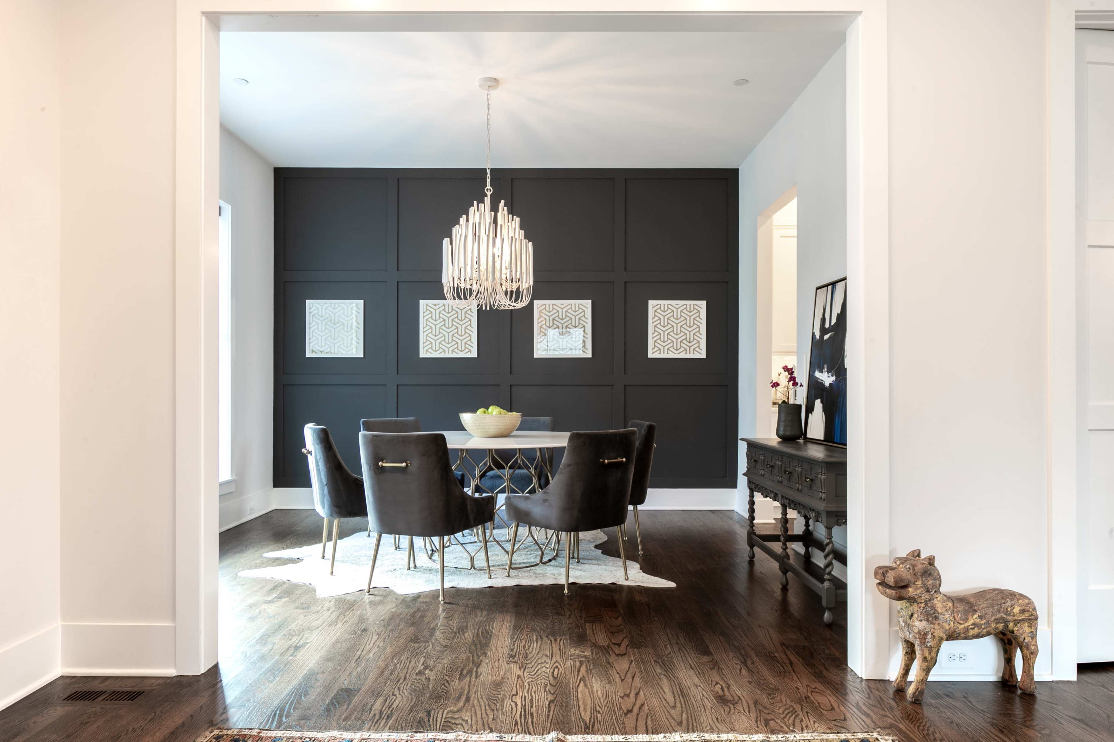 A dining room features a round table surrounded by upholstered chairs, a modern chandelier overhead, and dark accent walls with framed artwork.