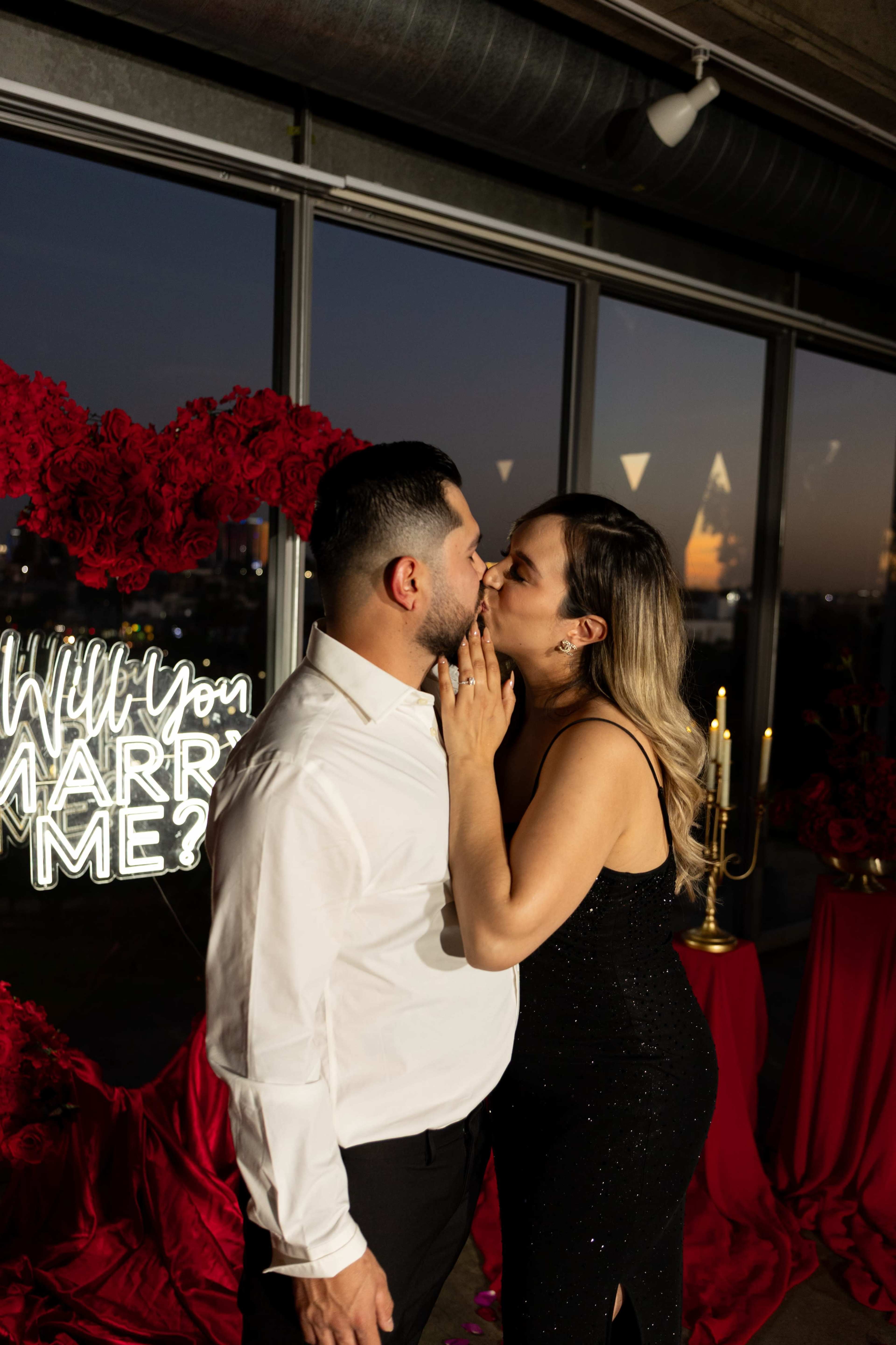 A couple embraces and kisses in front of a "Will You Marry Me?" backdrop adorned with roses and candlelight.