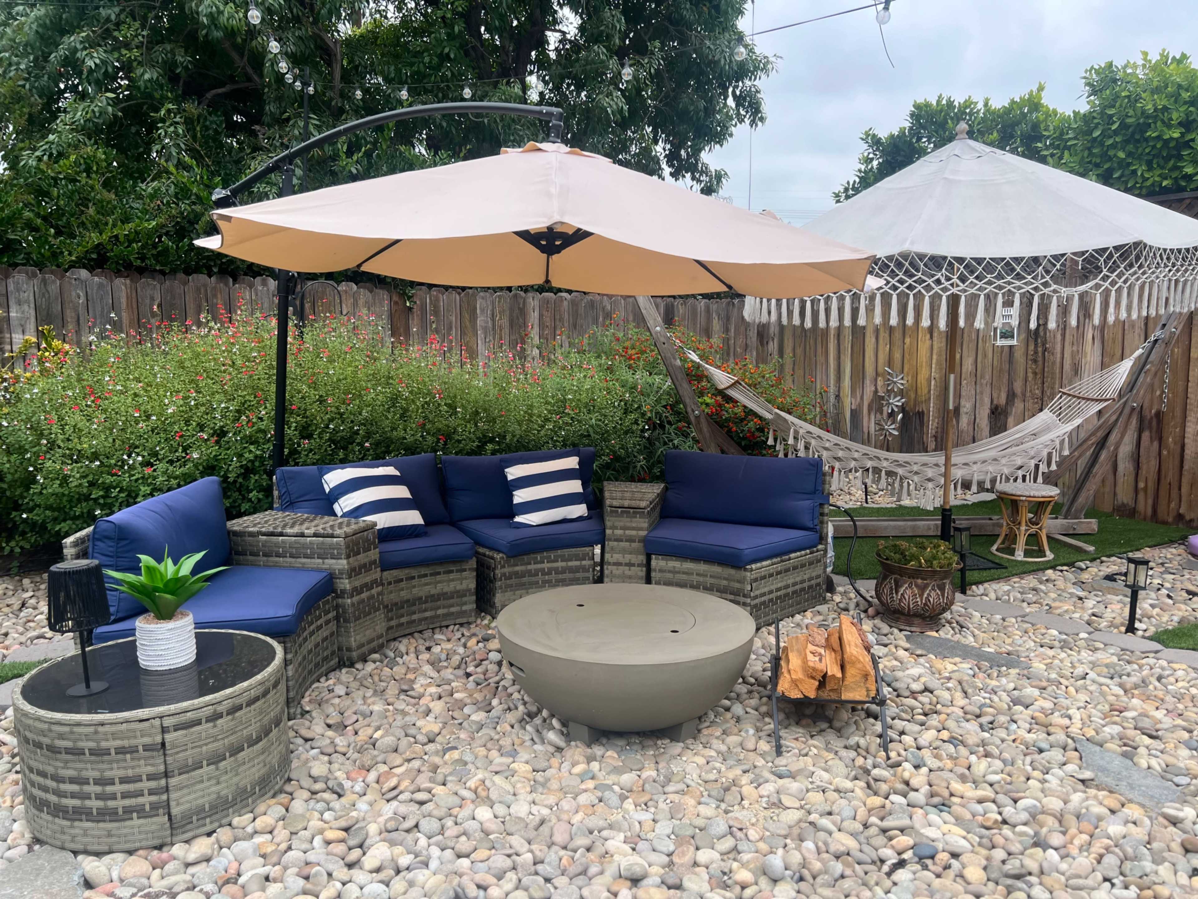 The image shows a cozy outdoor seating area with a curved sectional sofa, a round table, an umbrella, a hammock, and decorative plants set on a pebbled surface.