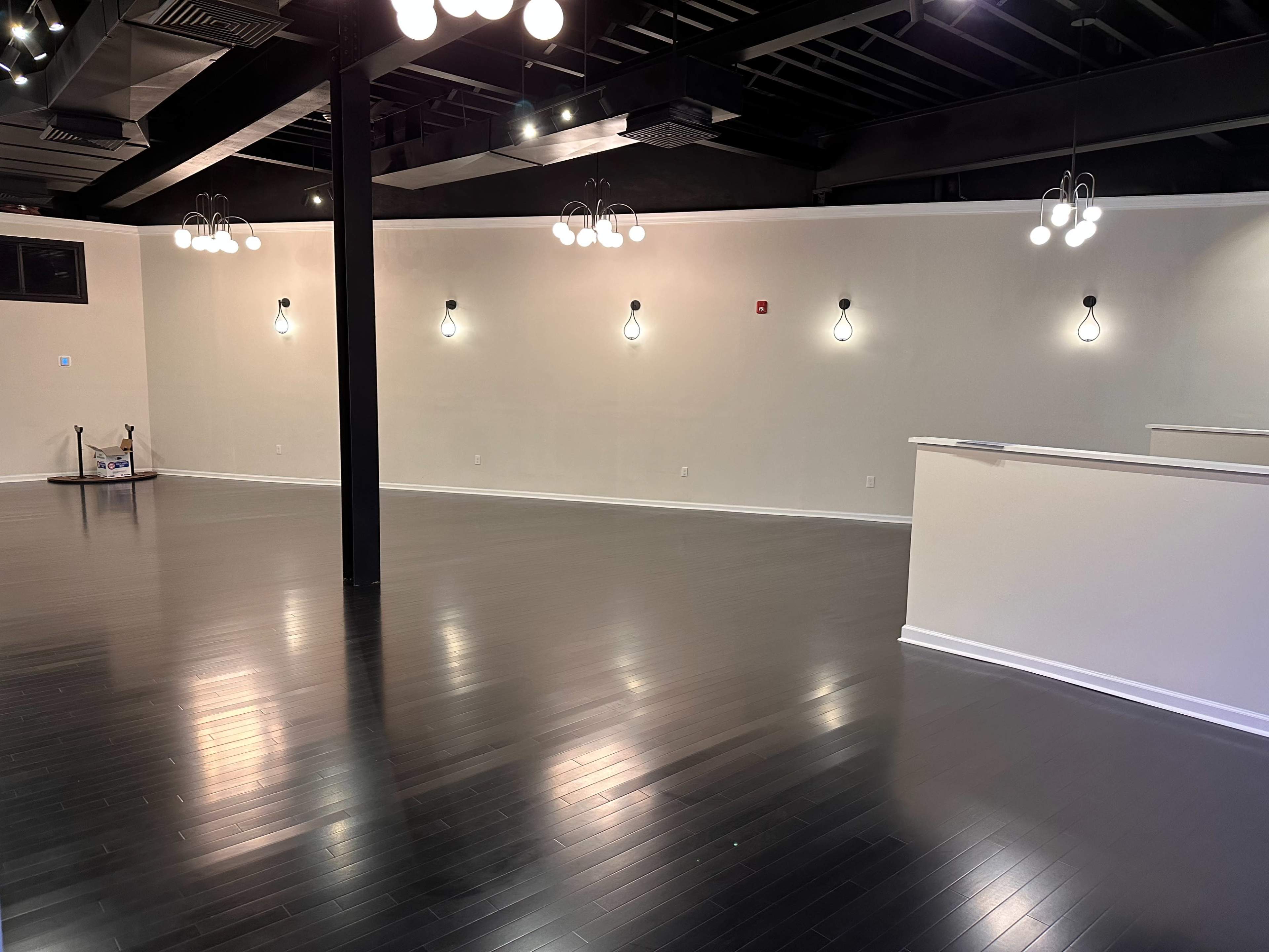 An empty interior space with polished dark wood flooring, white walls, and several light fixtures hanging from the ceiling.