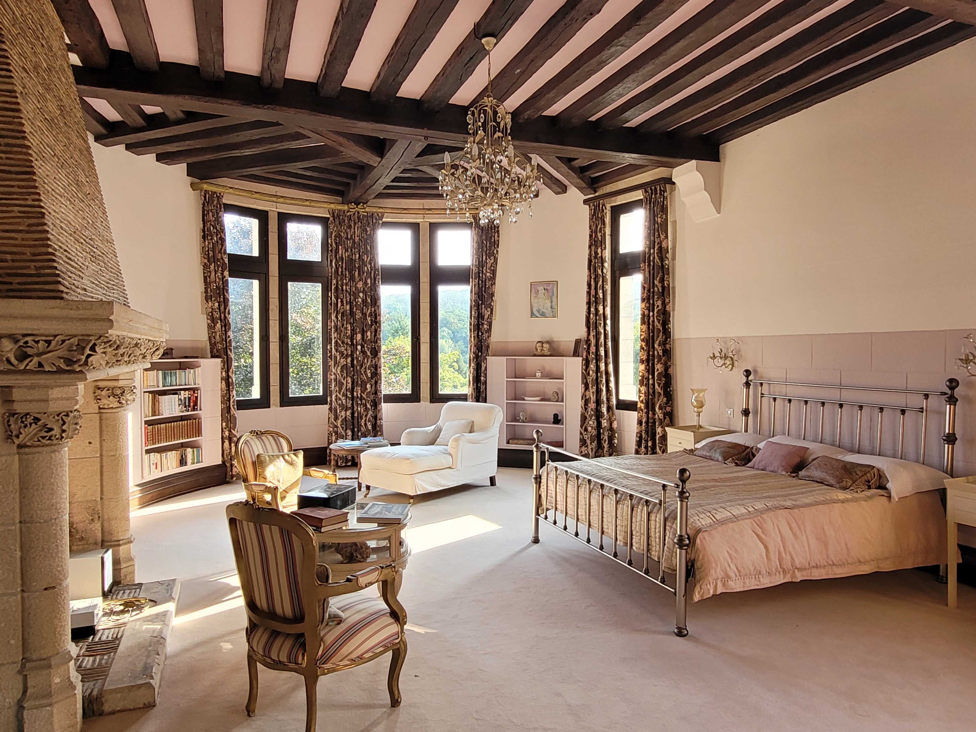 A spacious bedroom features a large bed, a cozy armchair, a fireplace, and large windows with views of the outside landscape.