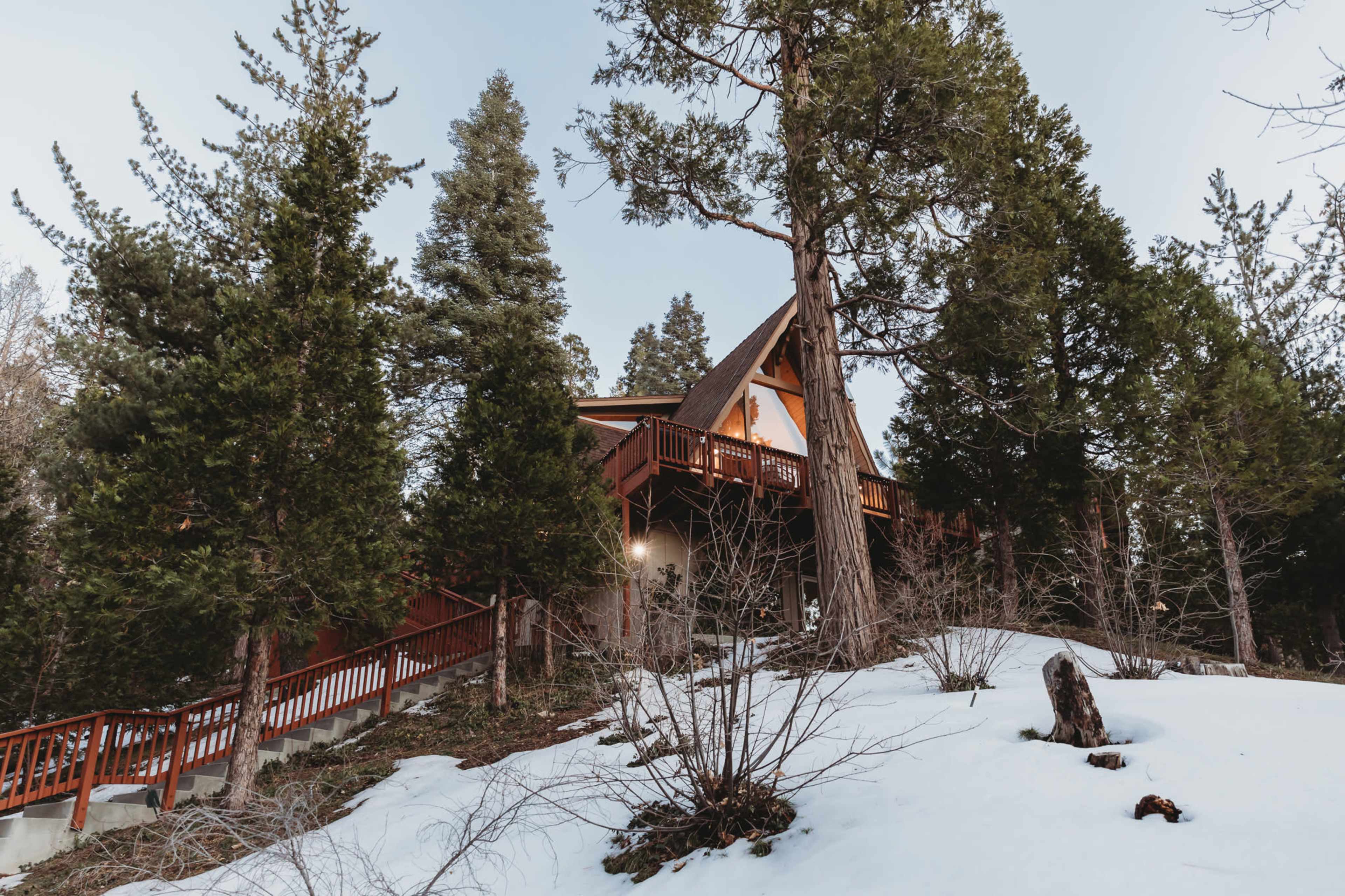 A wooden A-frame cabin sits among tall evergreen trees on a snowy landscape.