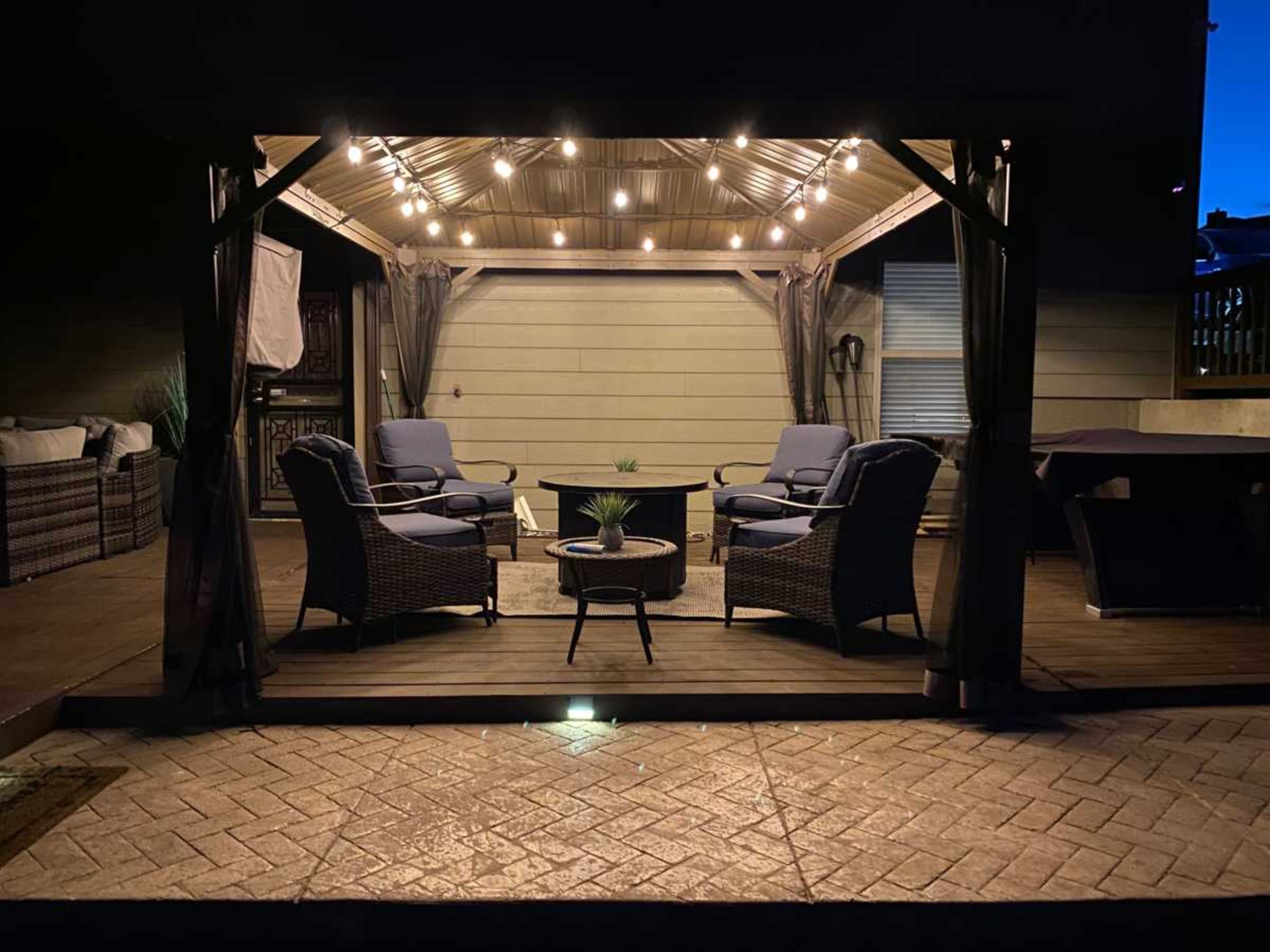 A covered outdoor seating area features two chairs and a table, illuminated by string lights against a twilight sky.