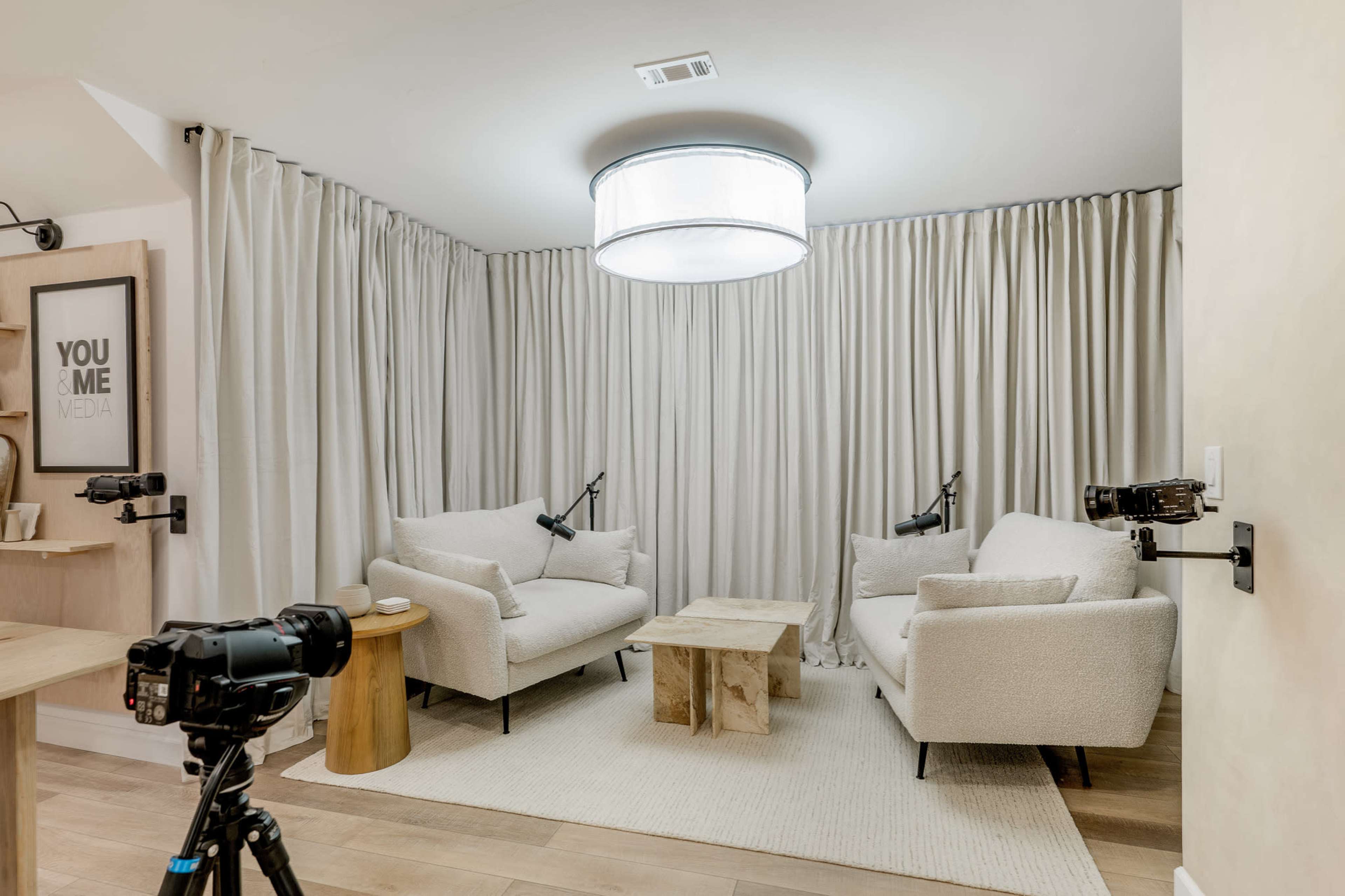A seating area with two light-colored armchairs facing a wooden coffee table, surrounded by beige curtains and equipped with two mounted cameras.