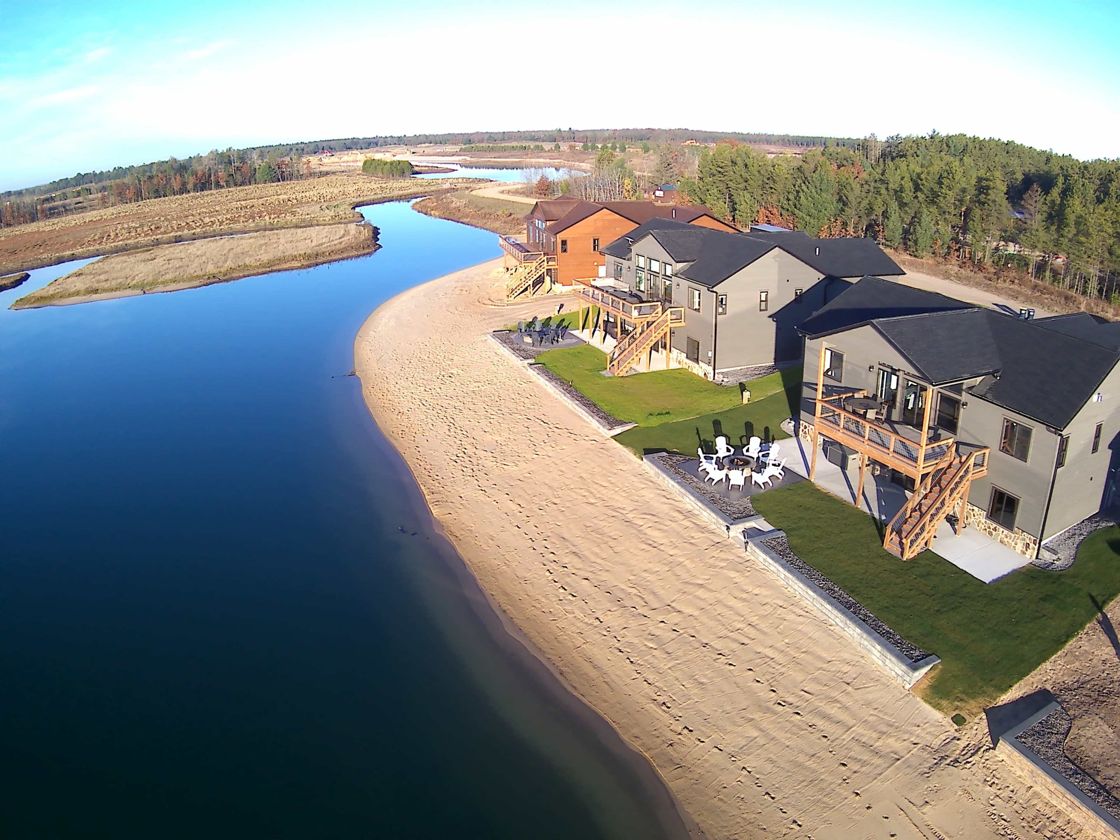 The image shows a shoreline with several houses overlooking a calm river, bordered by sandy beach and grassy areas.