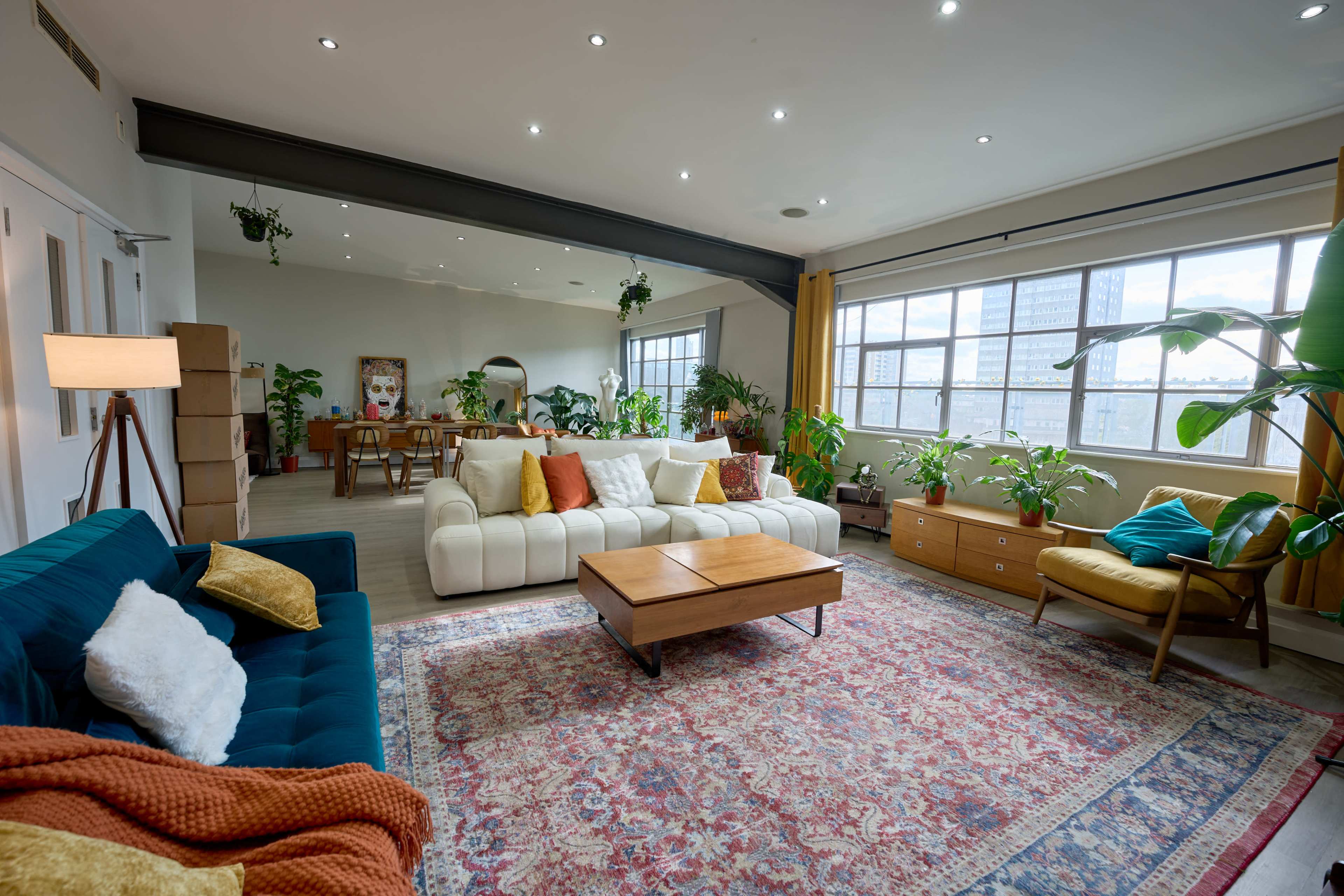 The image shows a modern living room with two sofas, a wooden coffee table, decorative plants, and large windows that let in natural light.