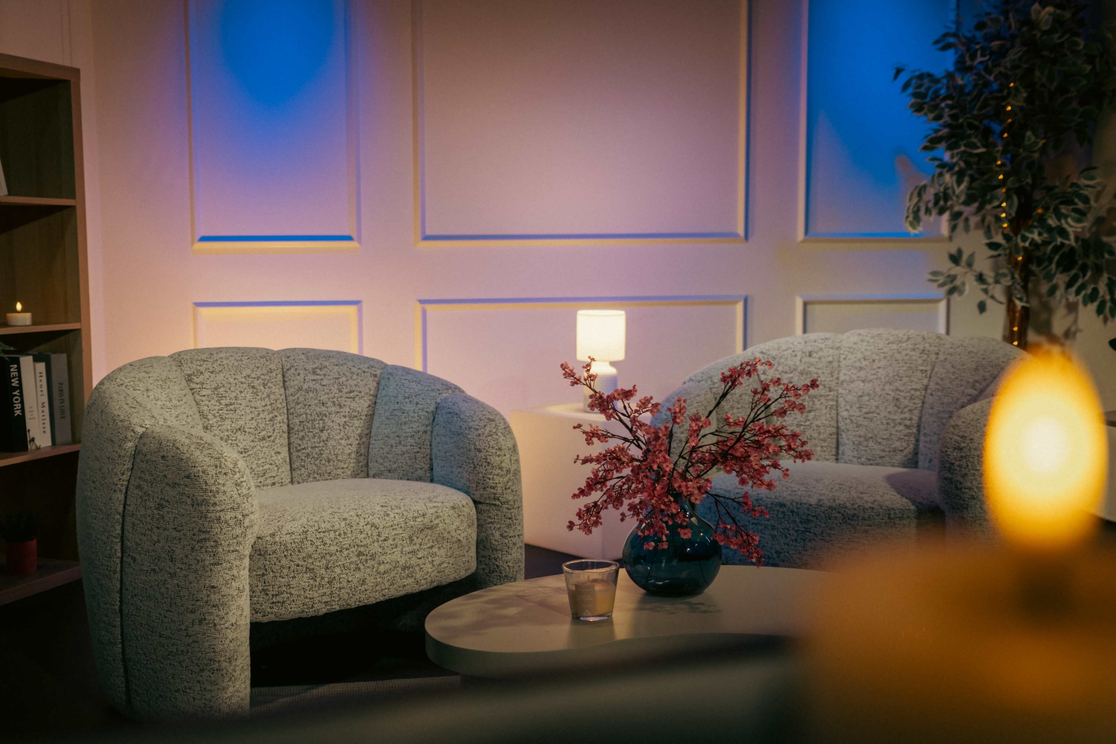 The image features a cozy indoor setting with two upholstered chairs, a small table, and a vase of flowers, illuminated by soft lighting.
