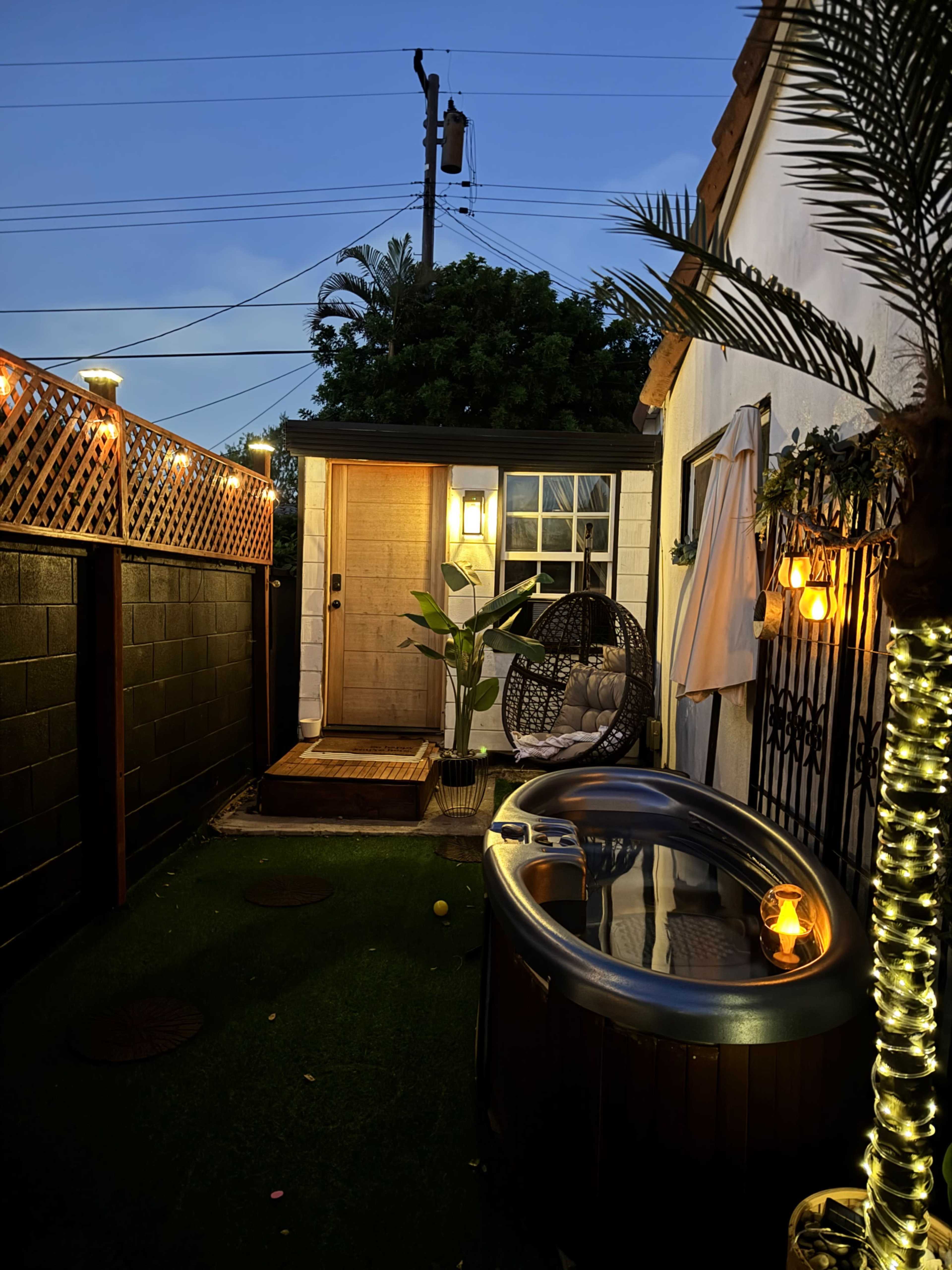 The image shows a cozy outdoor space featuring a hot tub, a hanging chair, and a door leading into a building, surrounded by decorative lighting and greenery.