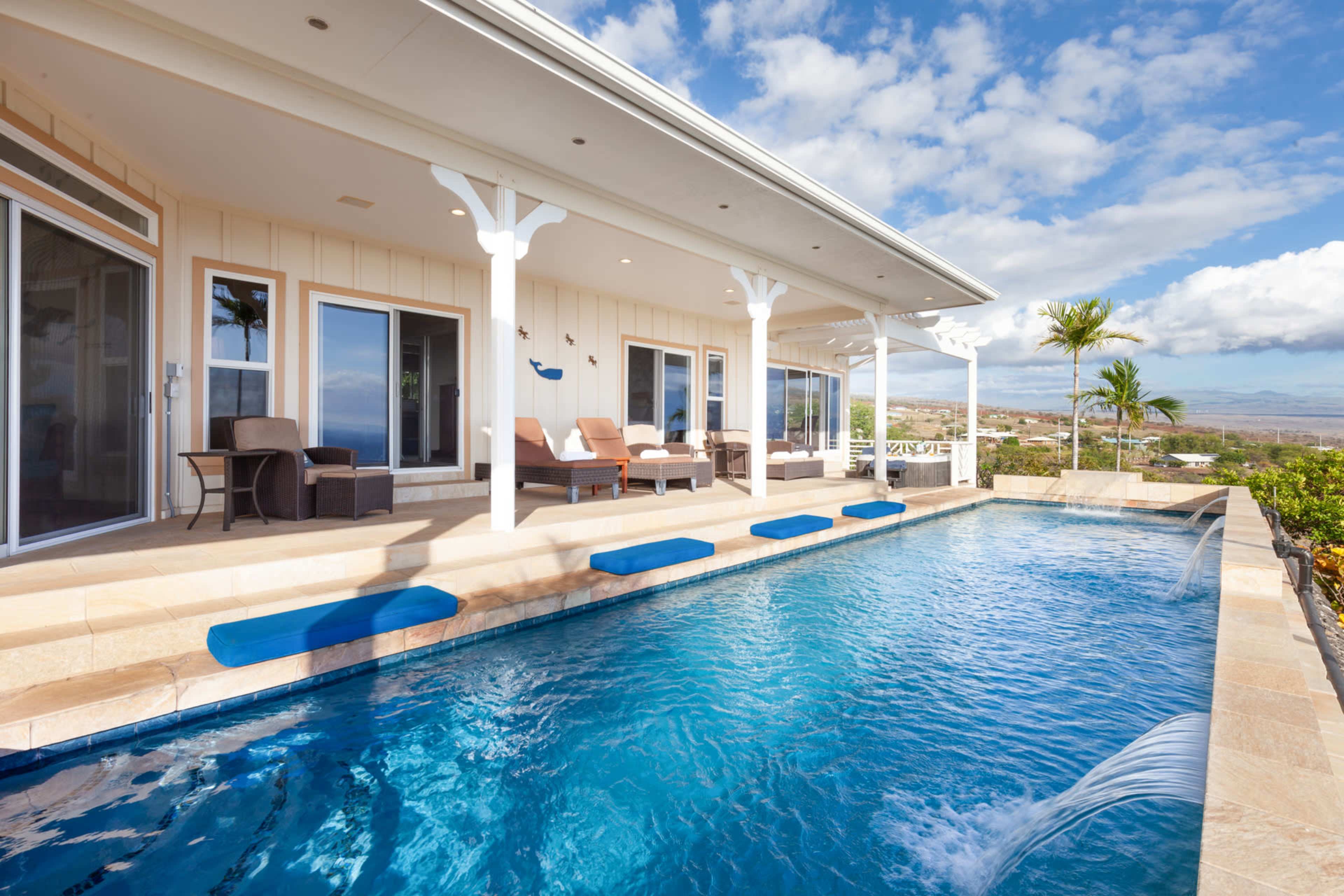 The image shows a modern house featuring a swimming pool with blue lounge mats, surrounded by tropical landscaping and clear skies.