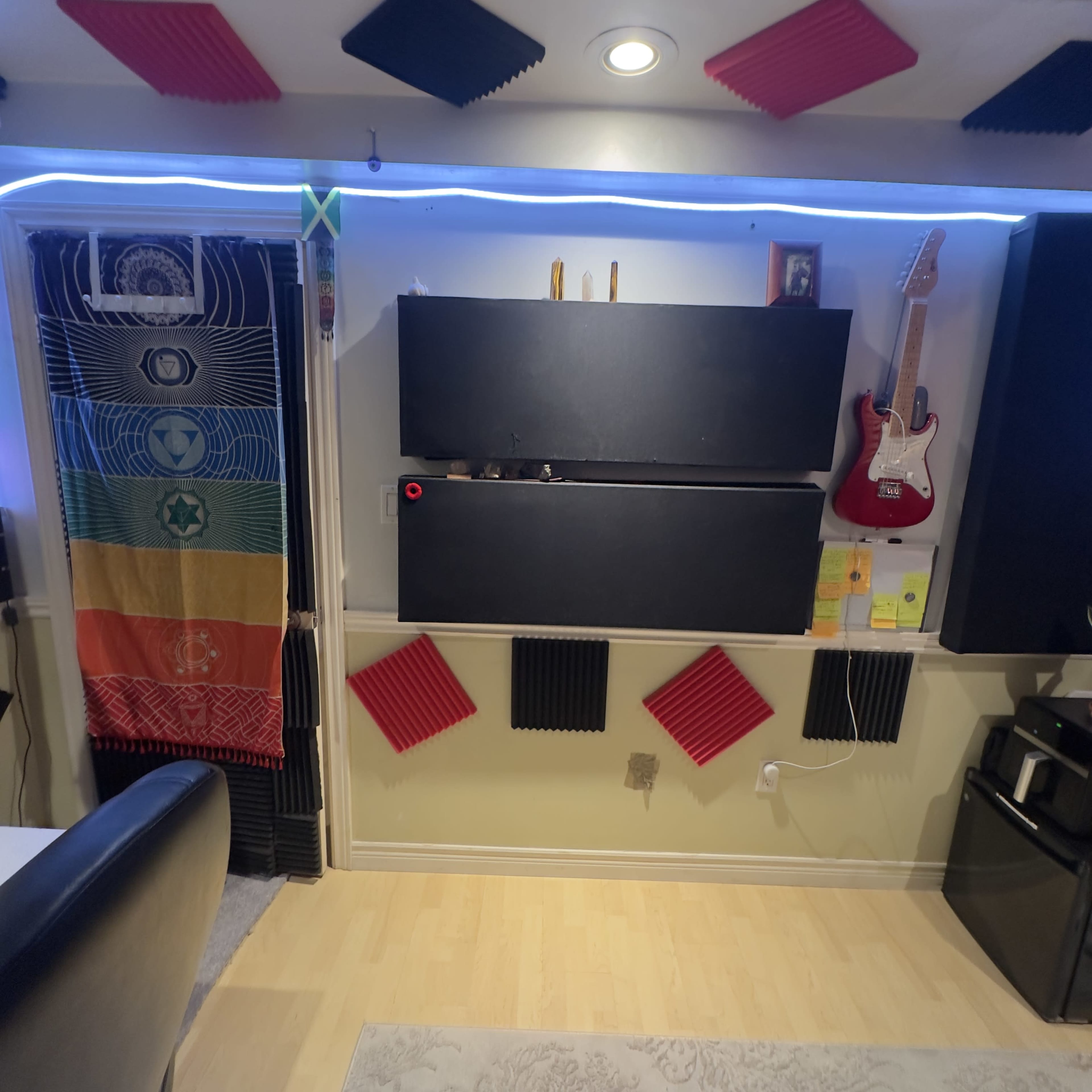 The image shows a room with soundproofing panels on the walls, a colorful banner, a guitar mounted on the wall, and storage boxes on a shelf.