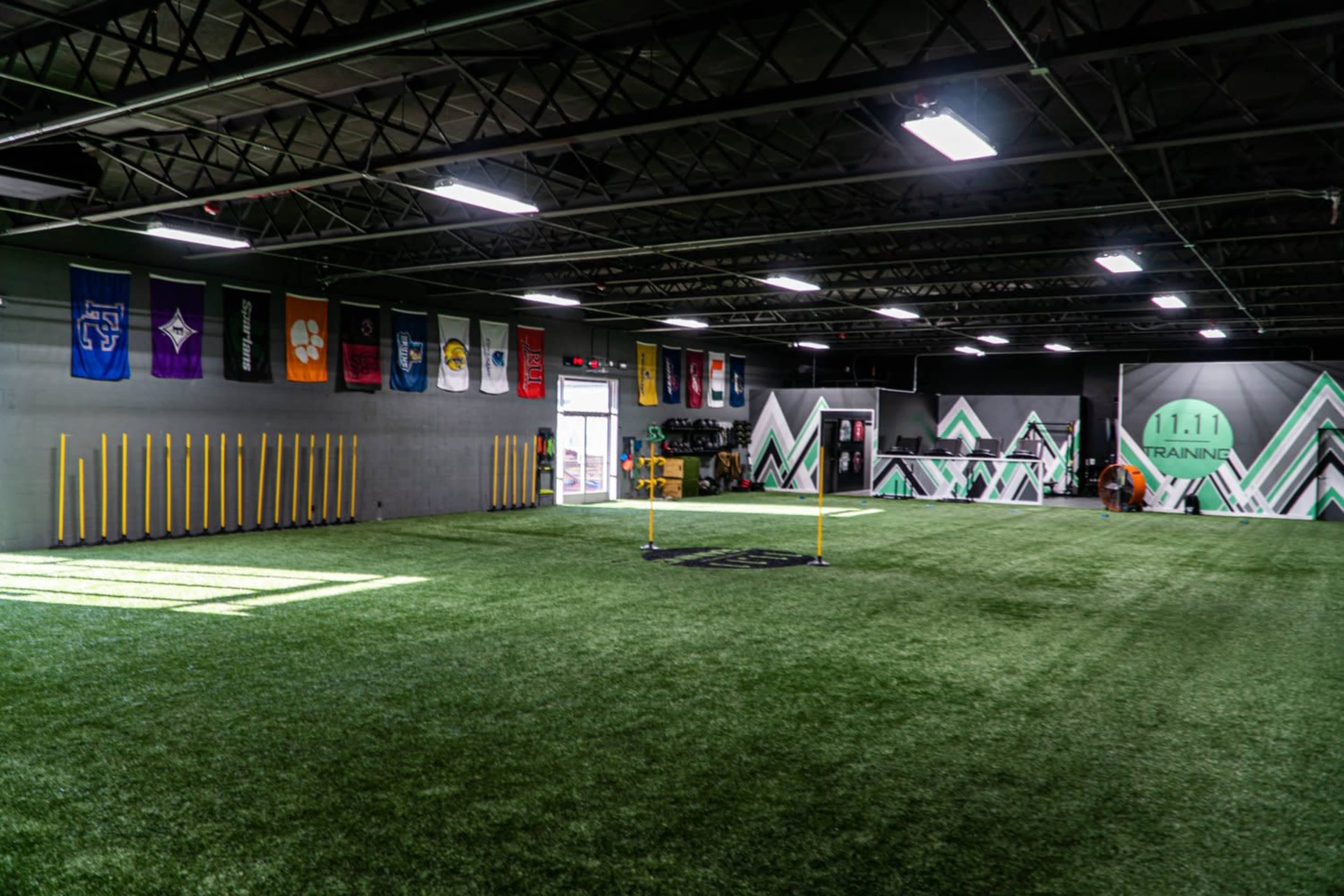 A spacious indoor training facility with artificial turf, featuring vertical yellow poles and various team banners on the walls.