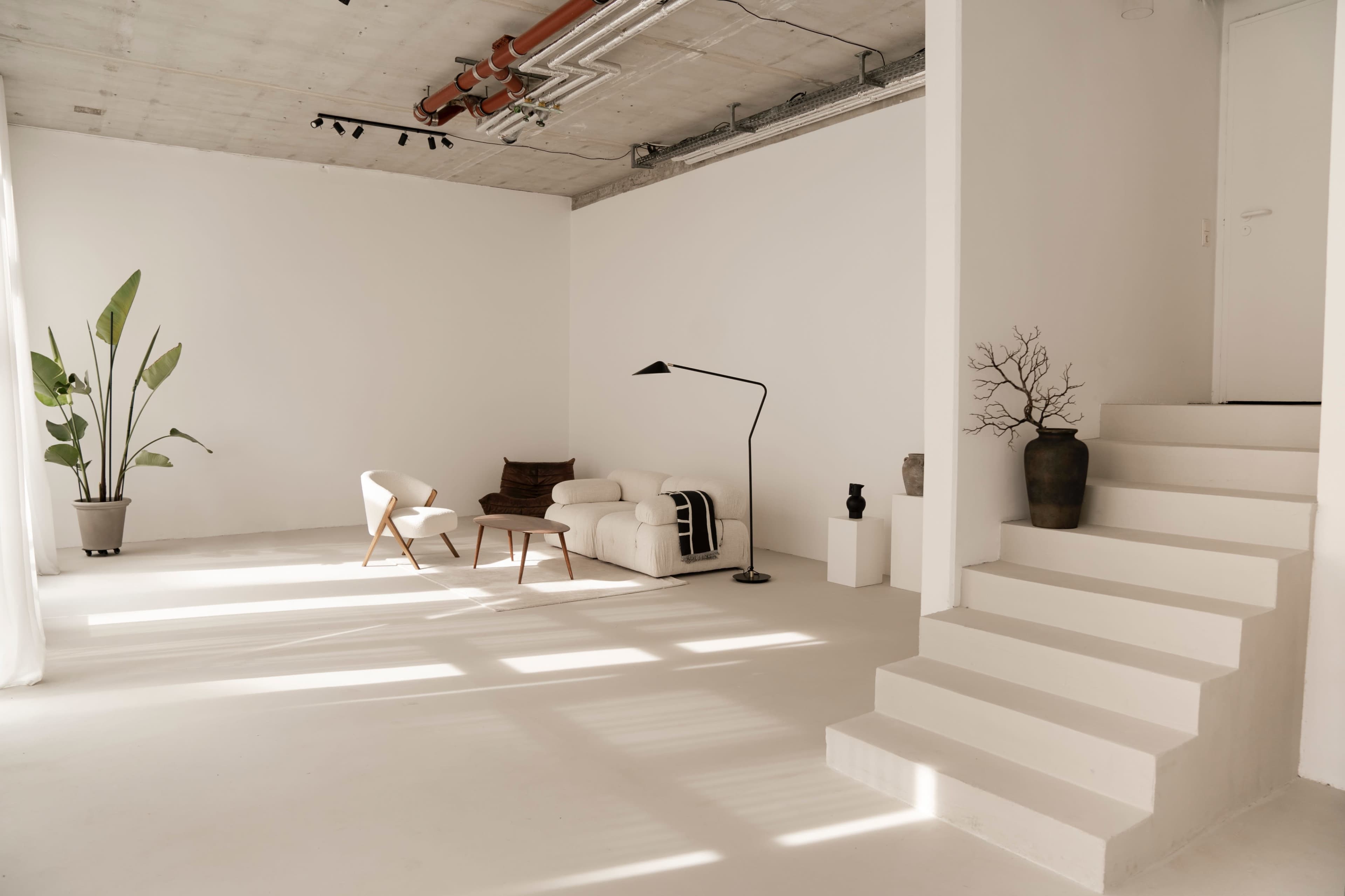 A bright, minimalist room features a white sofa, a chair, a floor lamp, and potted plants beside a staircase.