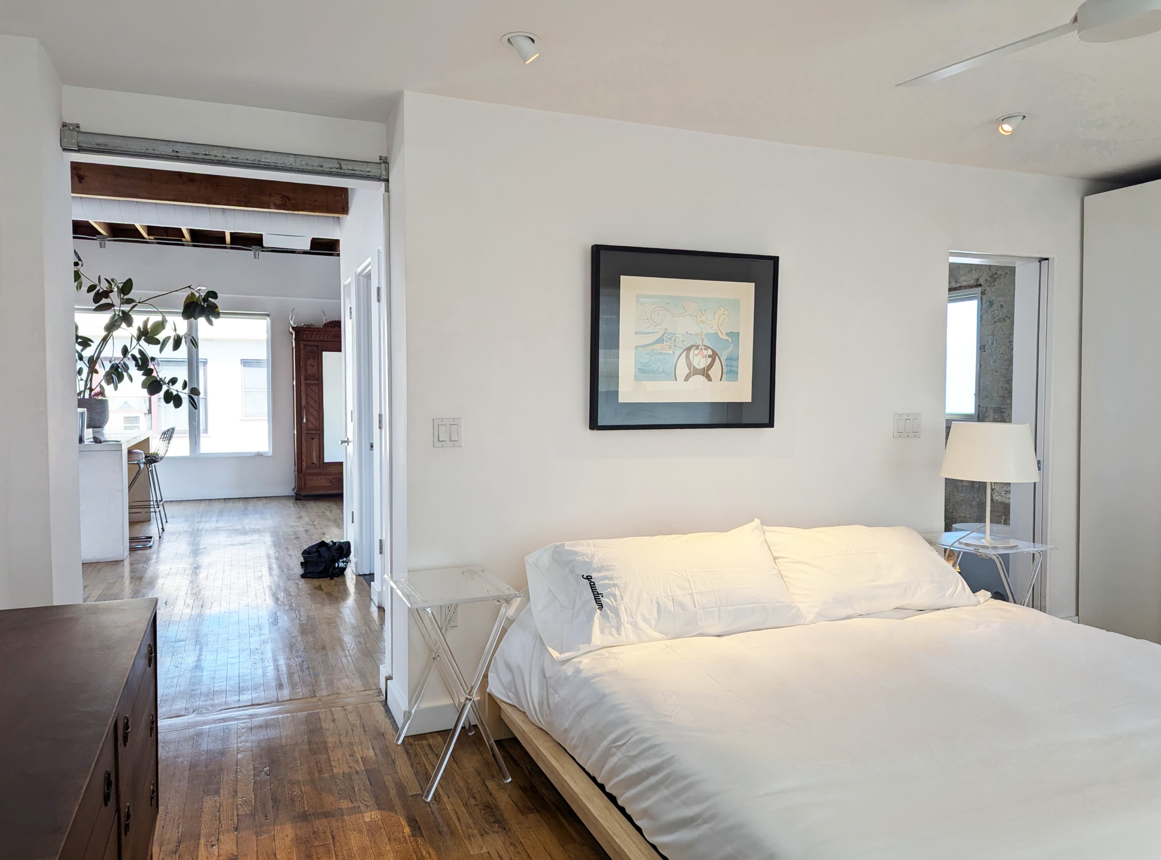 The image shows a minimalist bedroom with a bed, a bedside table, and a framed artwork on the wall, leading into a separate living space with large windows.