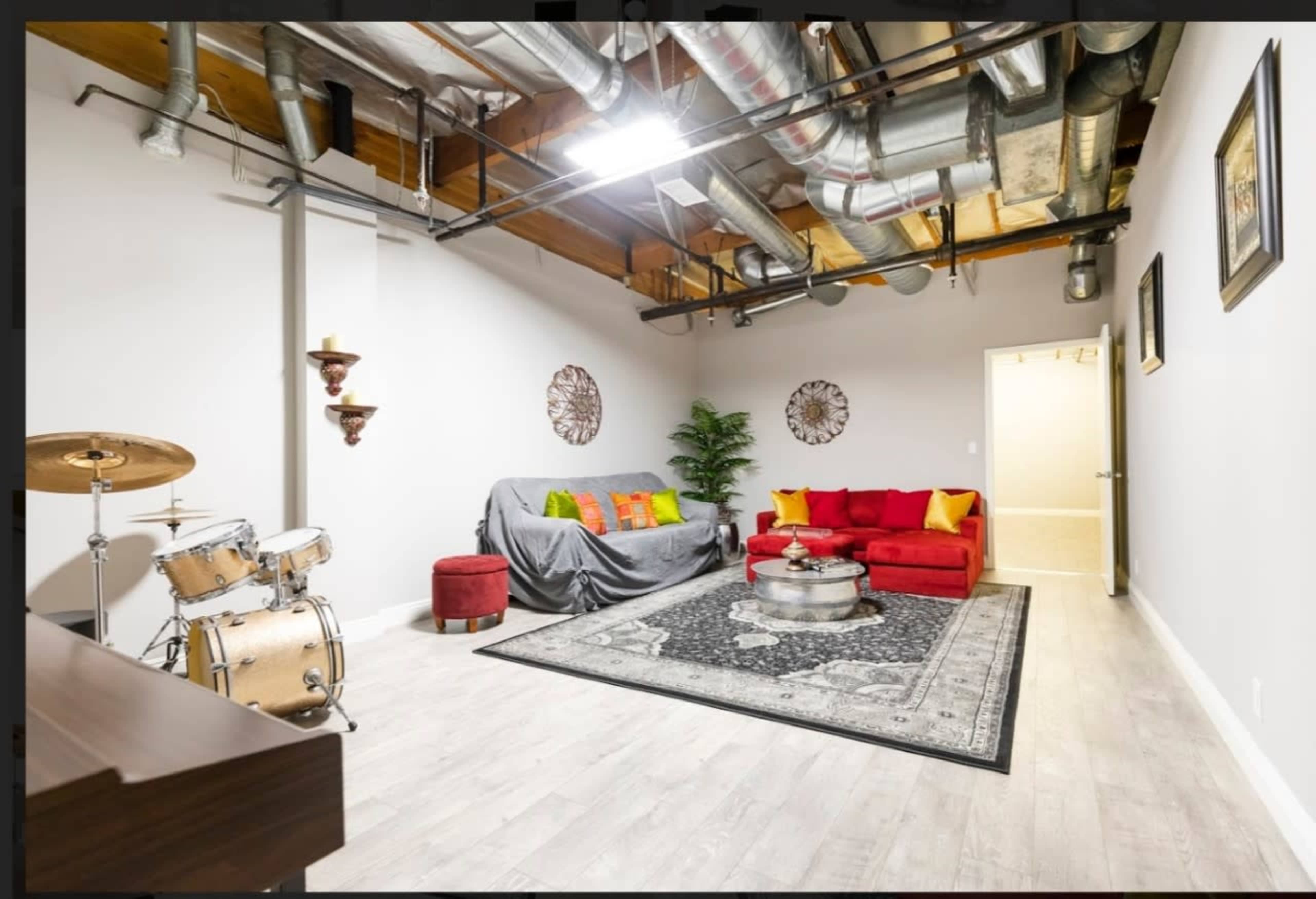 The room features a red sofa, a coffee table, a drum set, and decorative wall elements, with exposed metal ductwork overhead.