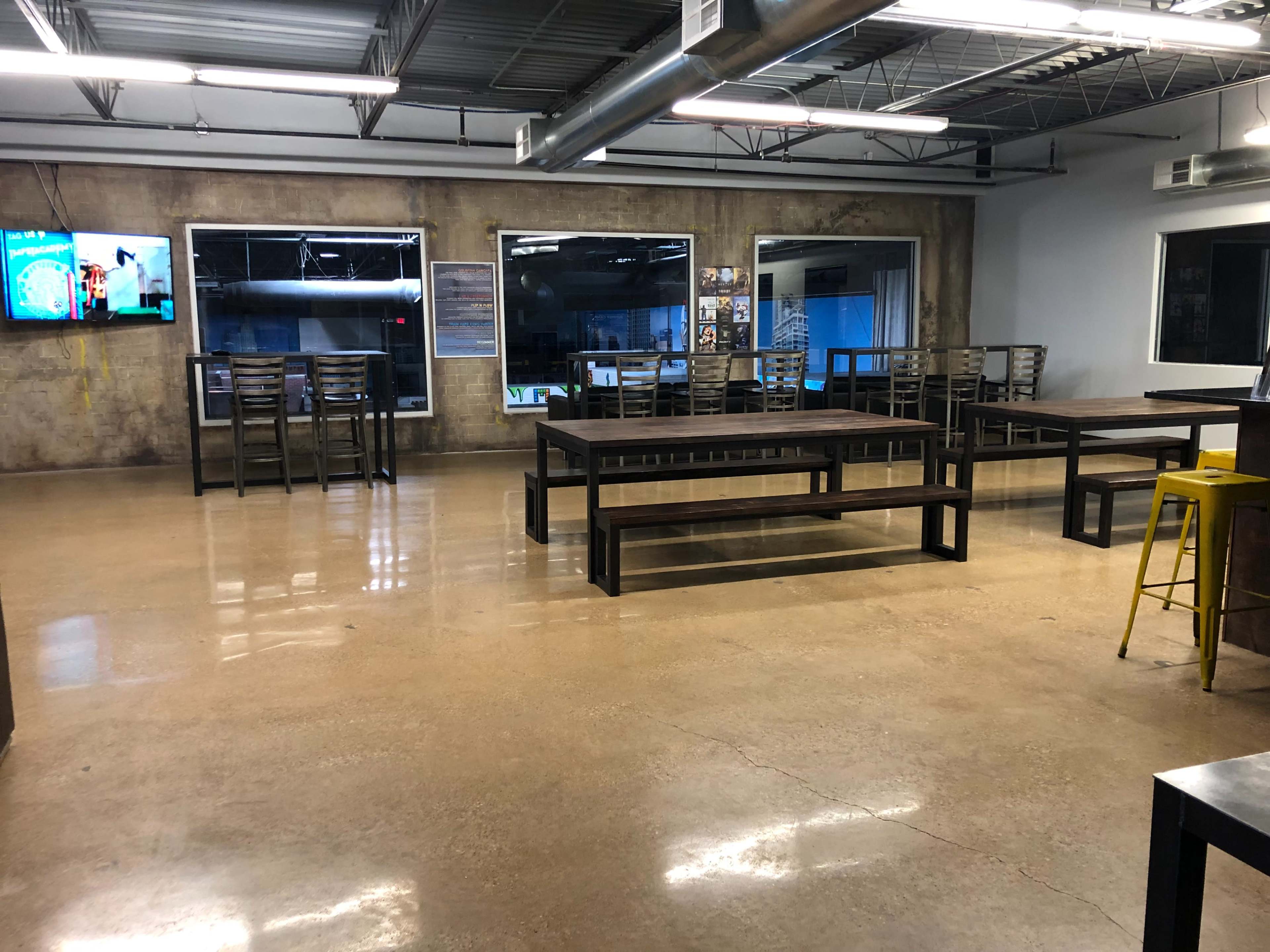 A spacious, industrial-style room with polished concrete floors, several tables, and large windows, featuring a television on one wall.