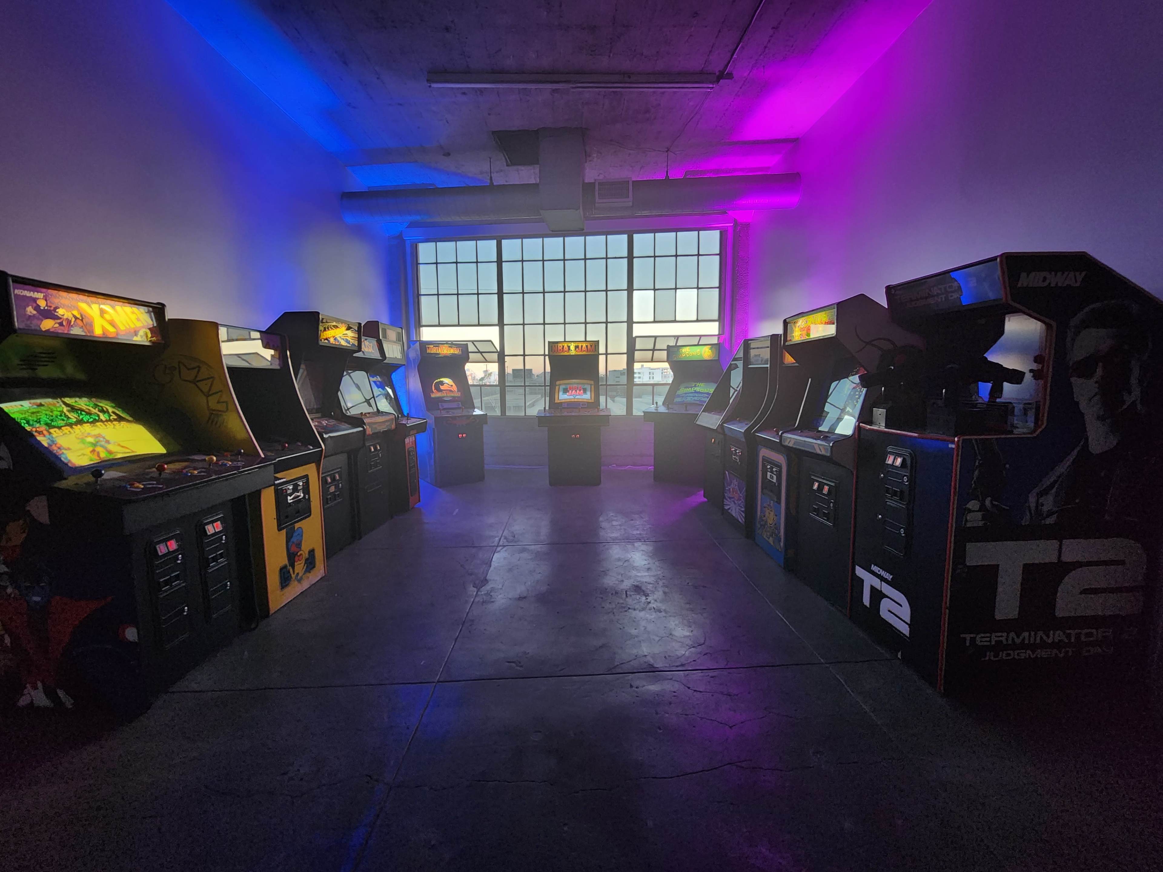 An arcade room with several vintage video game machines arranged in rows, illuminated by purple lighting near a large window.
