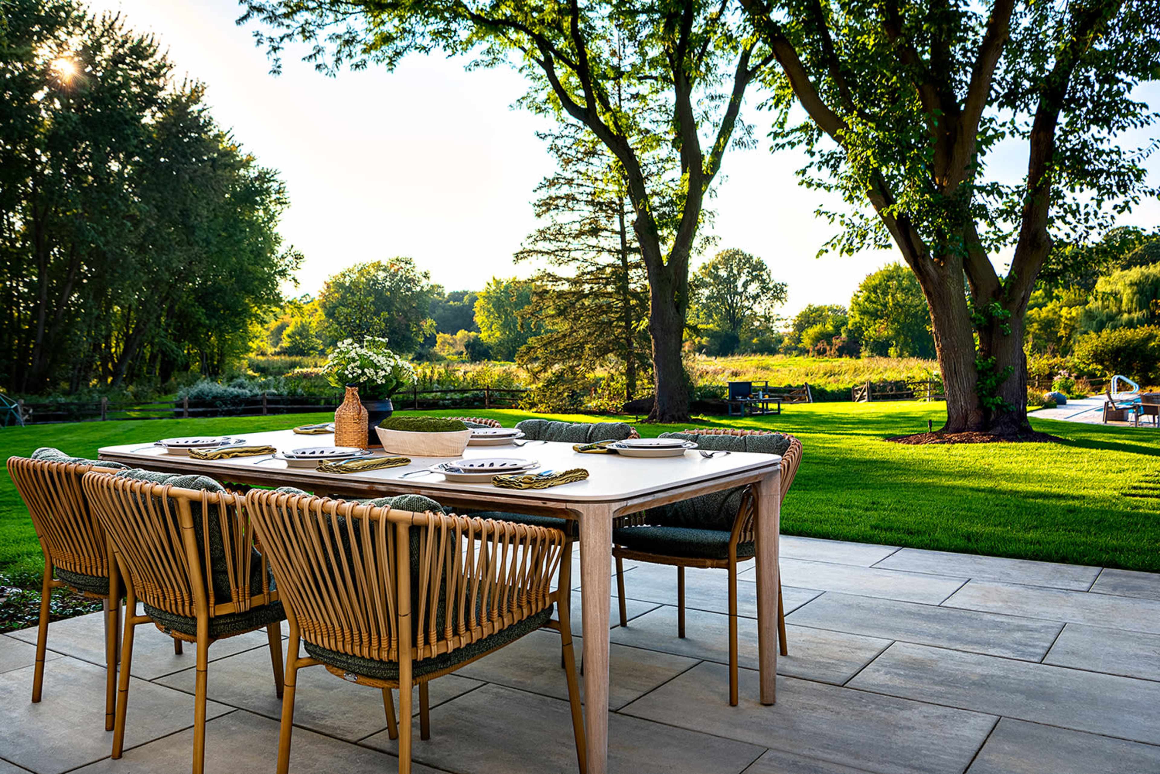 A wooden dining table is set with plates and decorative items on a patio surrounded by green grass and trees, with a view of a natural landscape in the background.