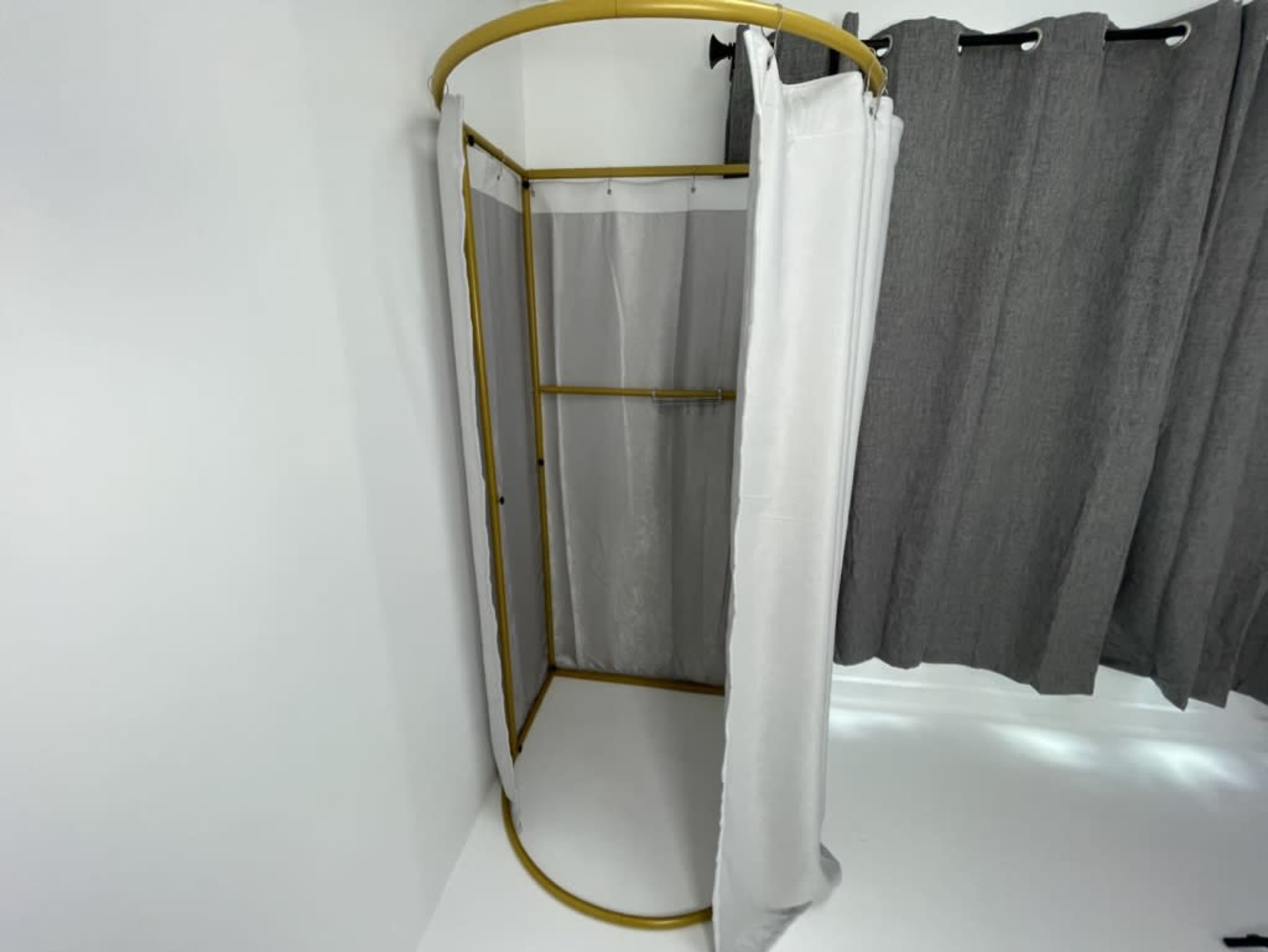 The image shows a minimalist shower area with a gold-framed structure and white curtains, situated in a corner with gray curtains nearby.