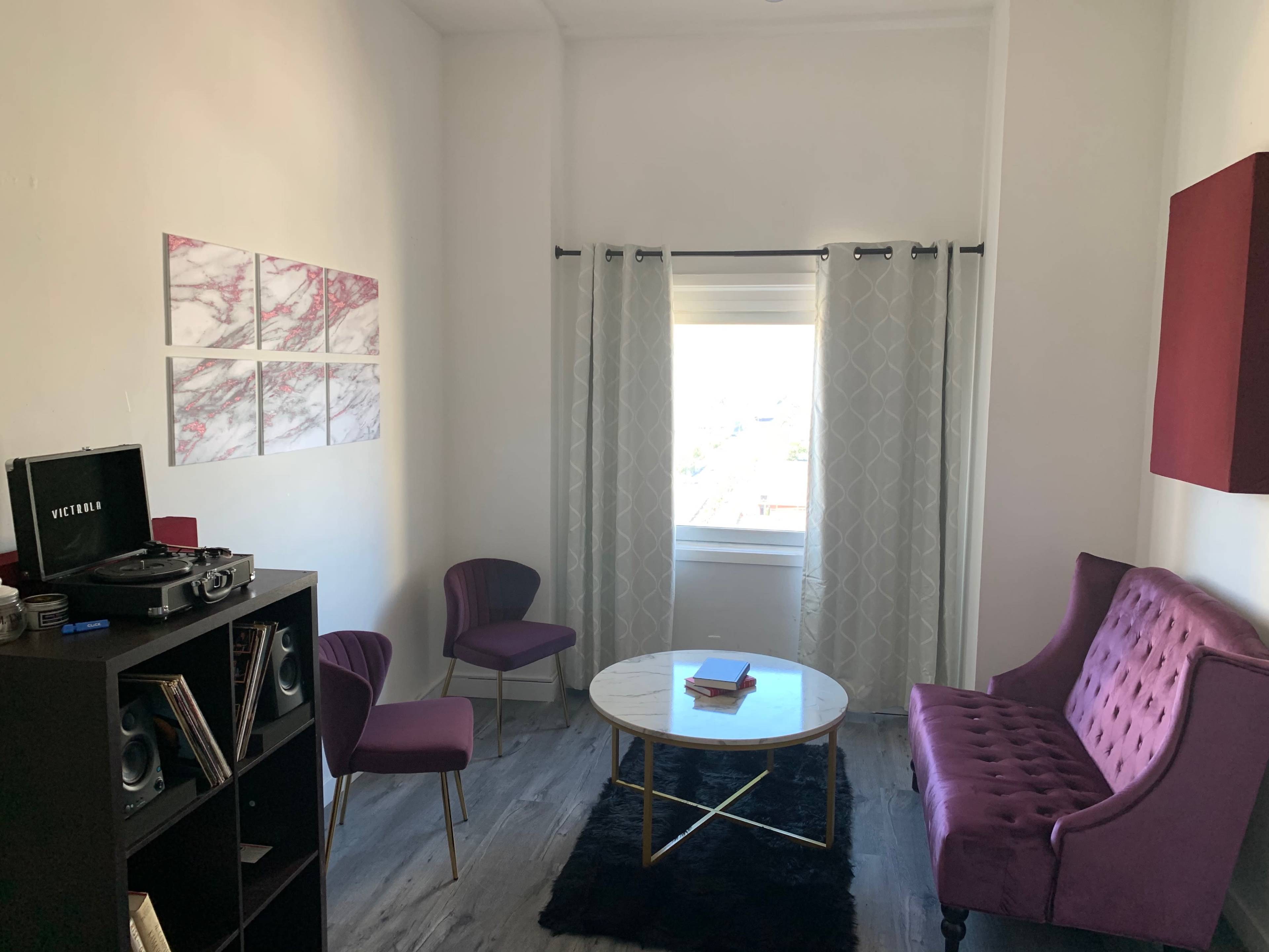 A small living room features a purple sofa, two chairs, a round coffee table, and a black bookshelf with a record player.