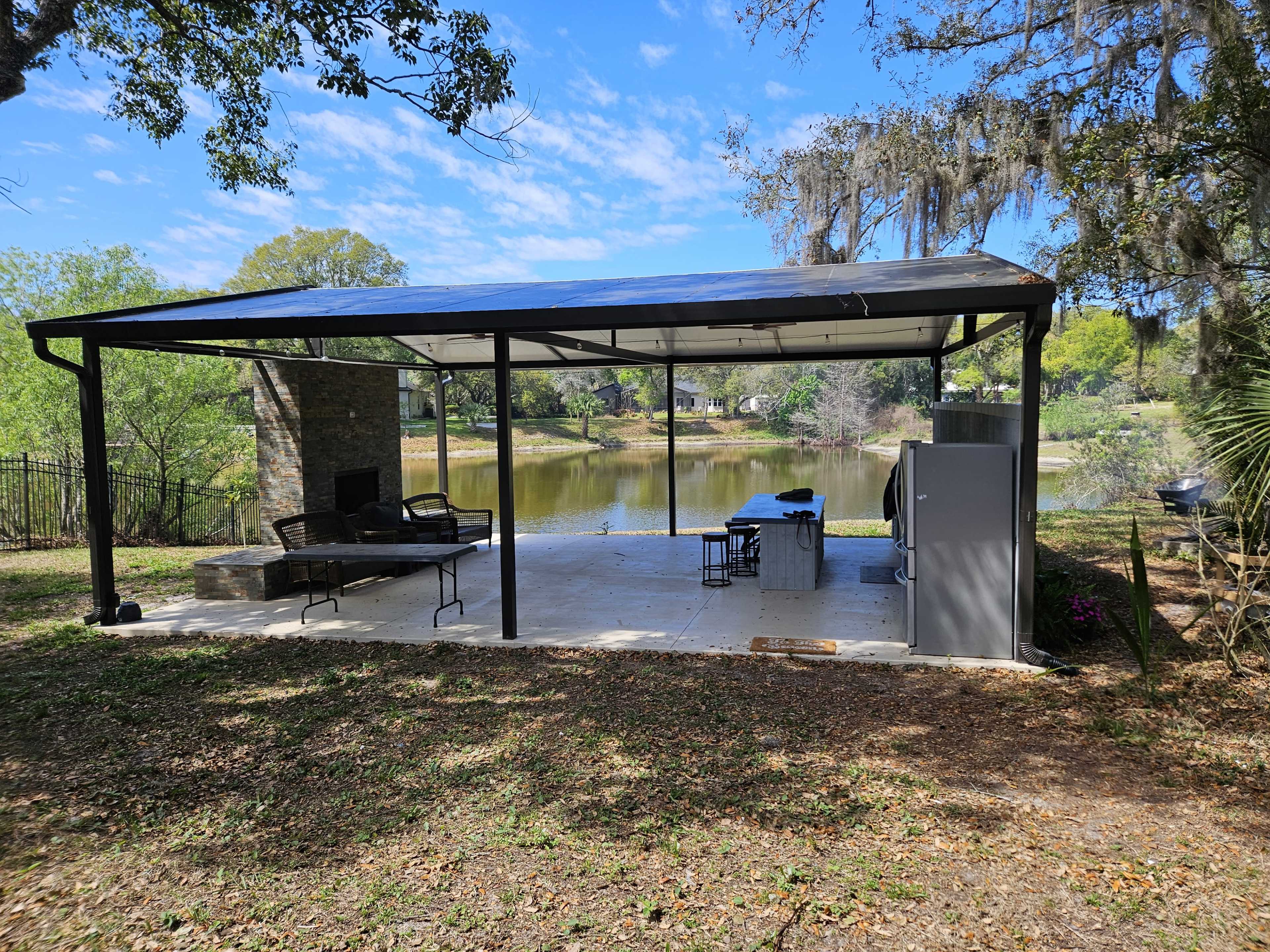 The image shows a covered outdoor patio with a seating area, a grill, and a view of a body of water surrounded by trees.