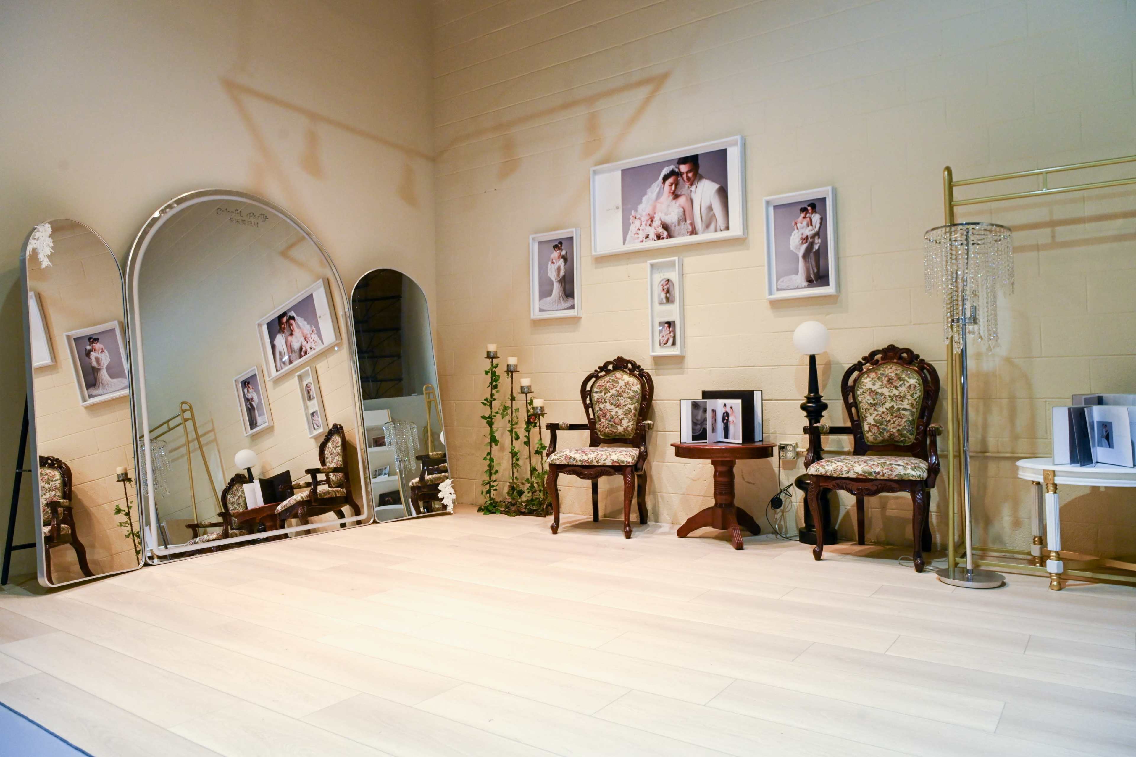 The image shows a stylish room featuring two ornate chairs, a small table, a mirror, decorative plants, and framed photographs on the walls.