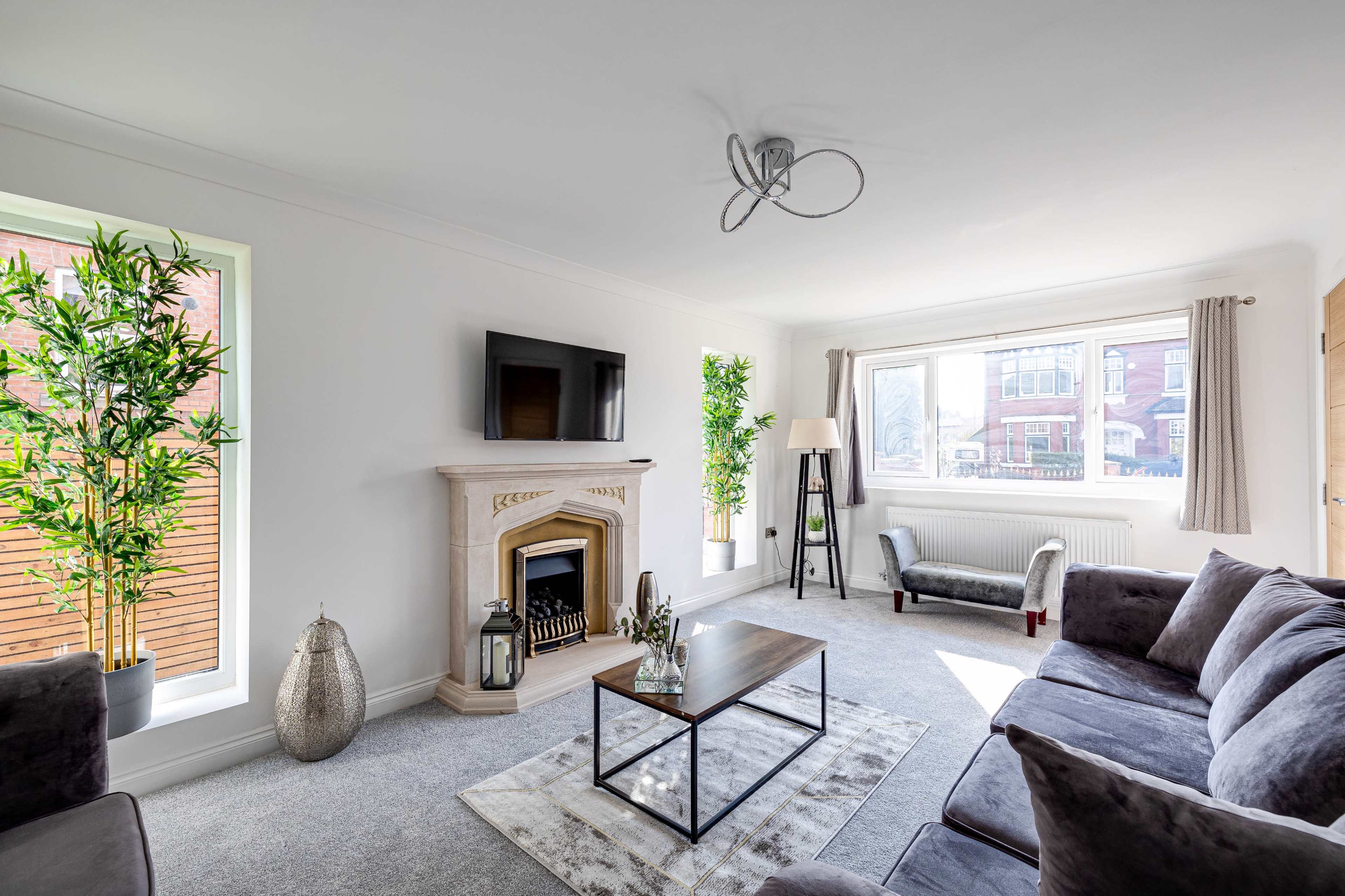 The living room features a stone fireplace, a modern television mounted on the wall, a coffee table in the center, and large windows with plants on either side.