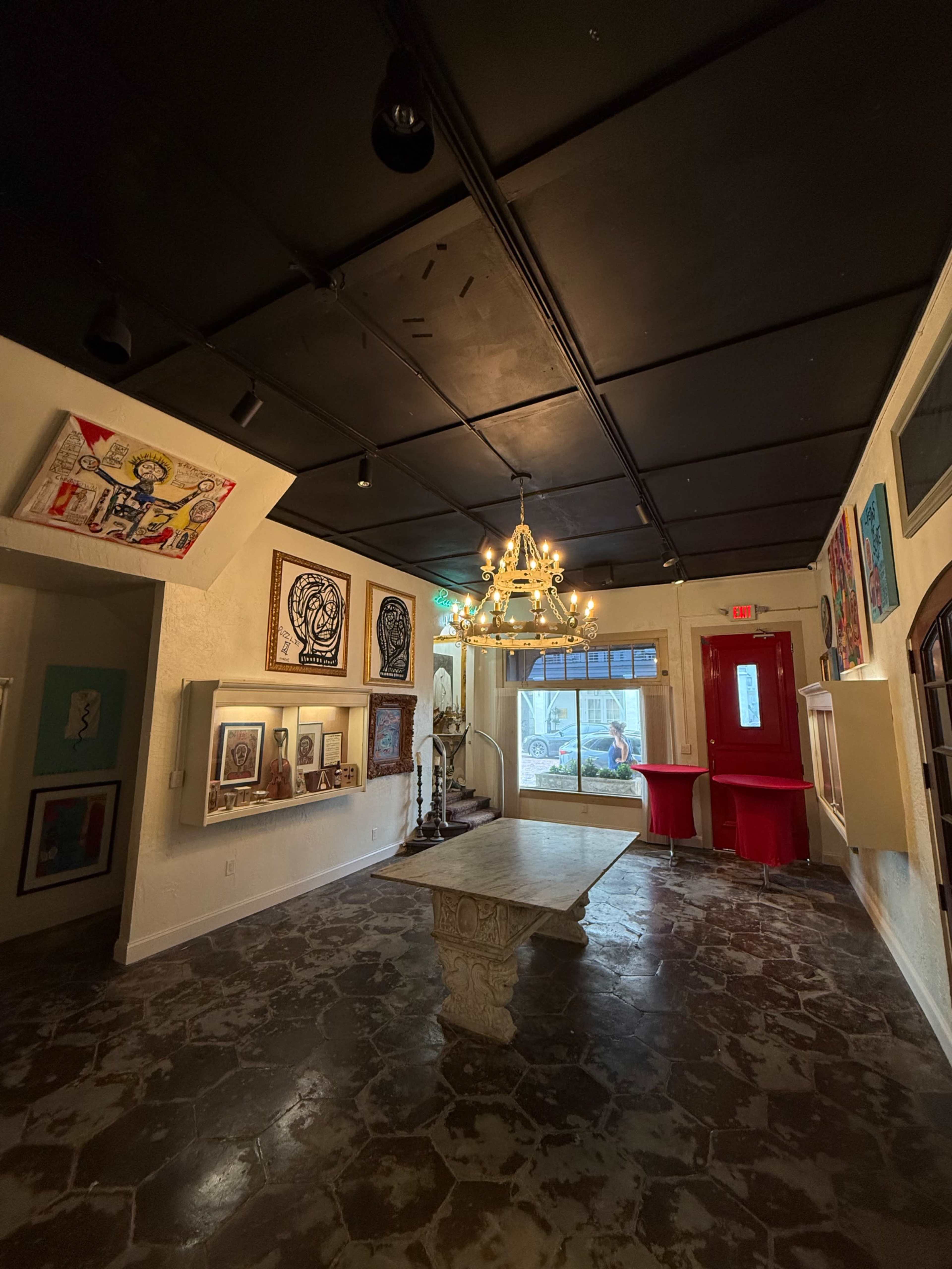 The image shows an art gallery interior featuring various framed artworks on the walls, a central stone table, and a chandelier hanging from the ceiling.