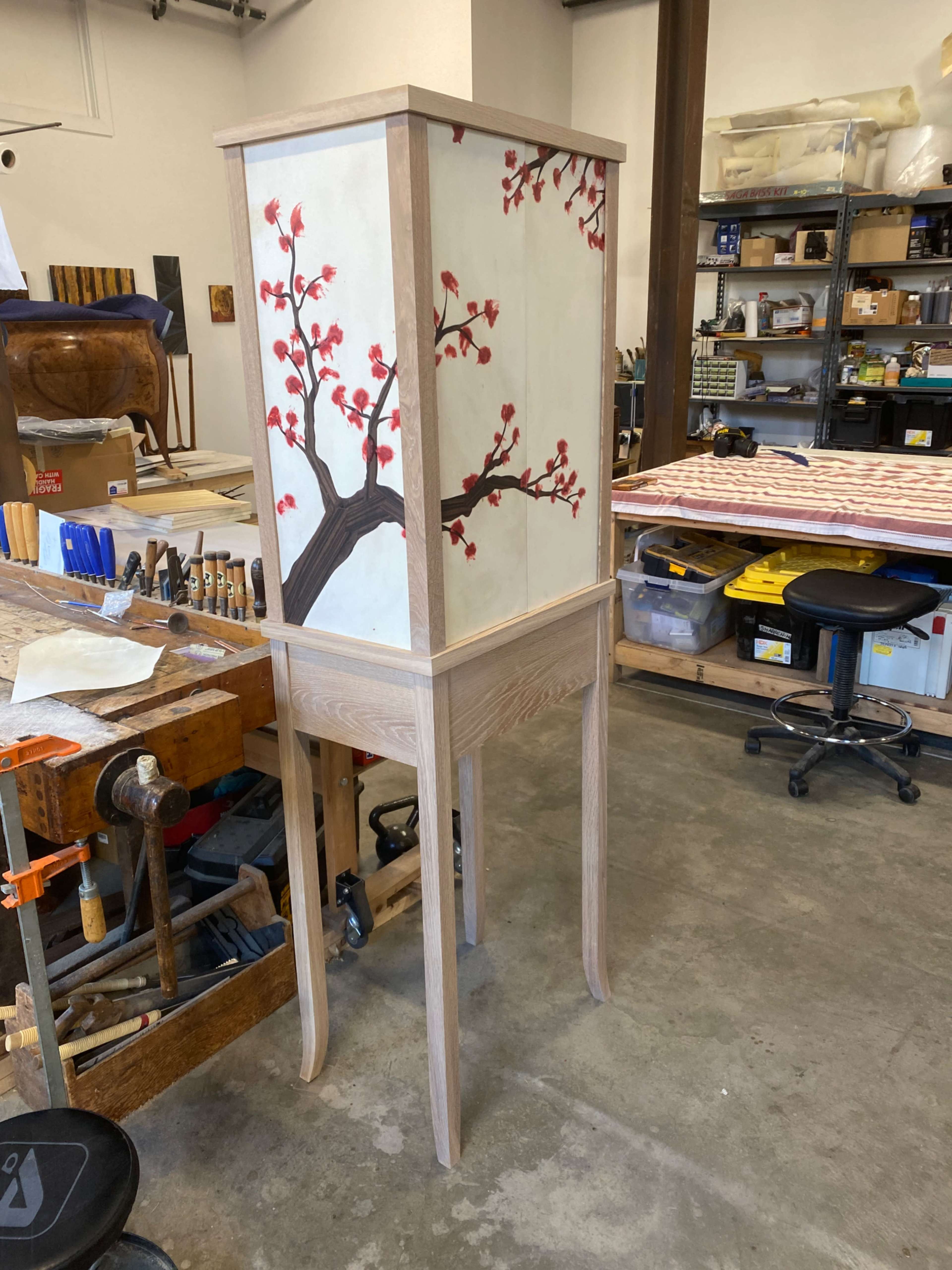 The image shows a tall wooden cabinet with a decorative design of a tree branch and blossoms on its sides, located in a workshop filled with various tools and materials.
