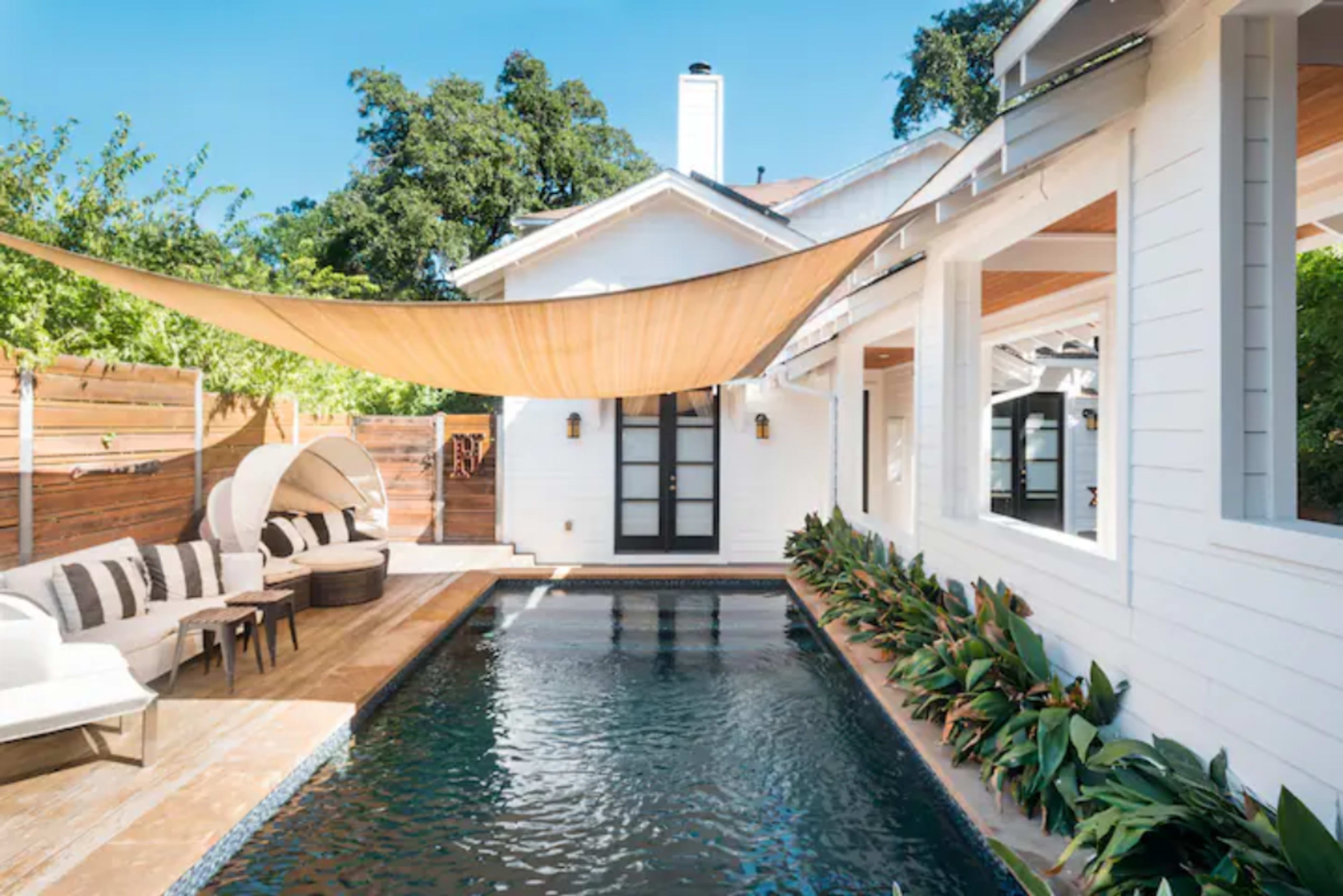 The image shows a modern outdoor living space featuring a rectangular swimming pool surrounded by plants, a cozy seating area, and a large shade sail overhead.