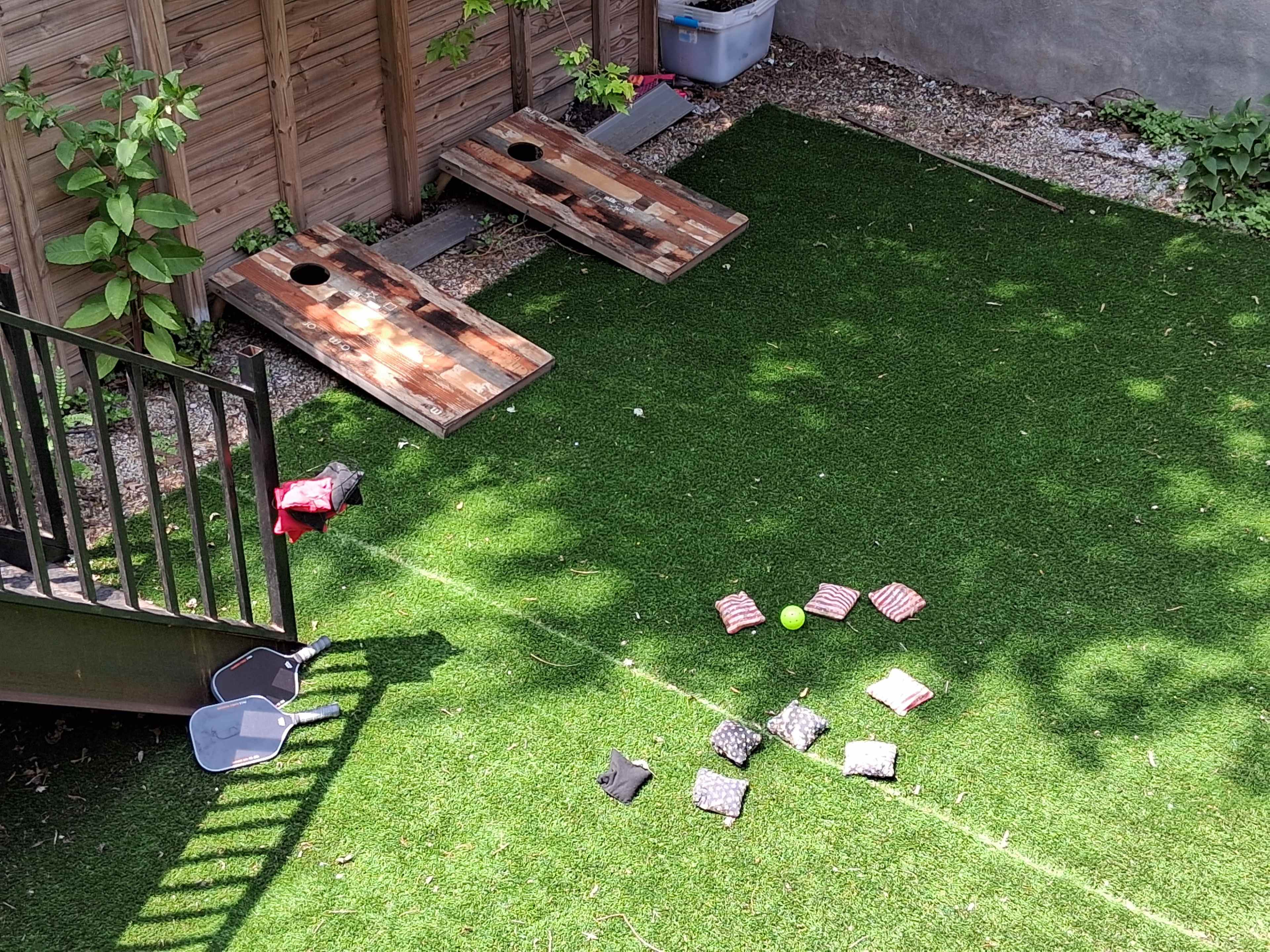 The image shows a backyard with two wooden cornhole boards set up on artificial grass, alongside scattered bags and a few objects on the ground.