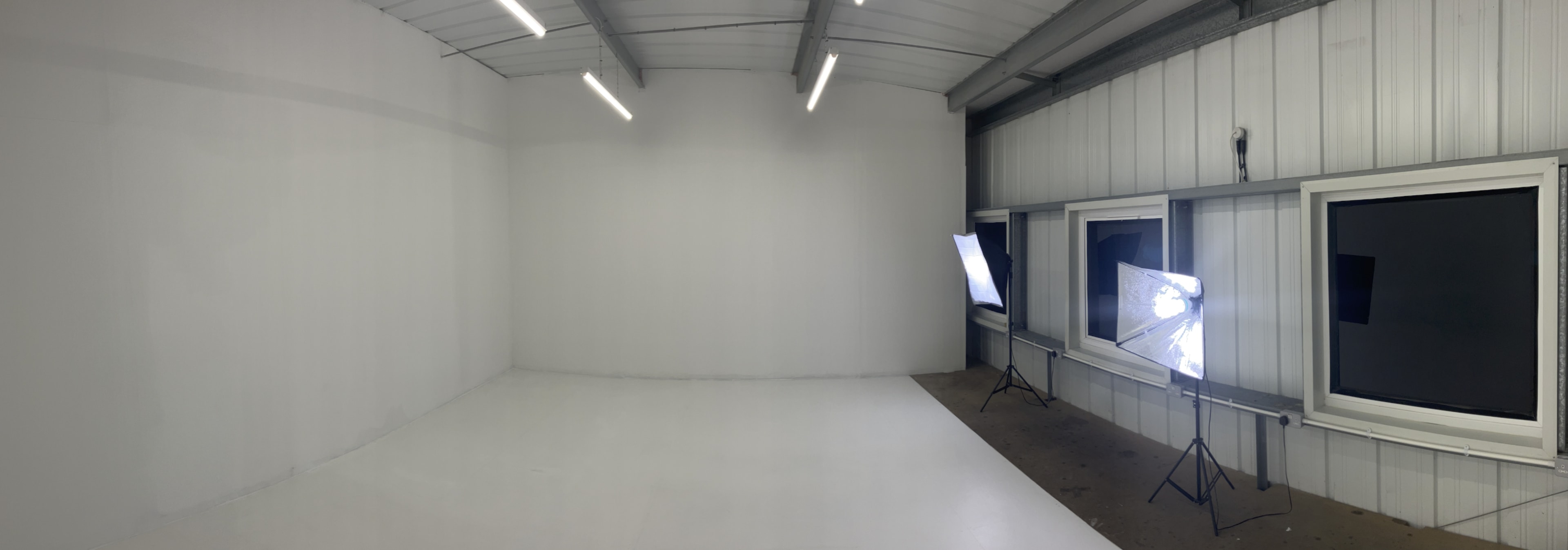 The image shows a spacious, blank studio environment featuring white walls, a white floor, and two windows with lighting equipment positioned outside.