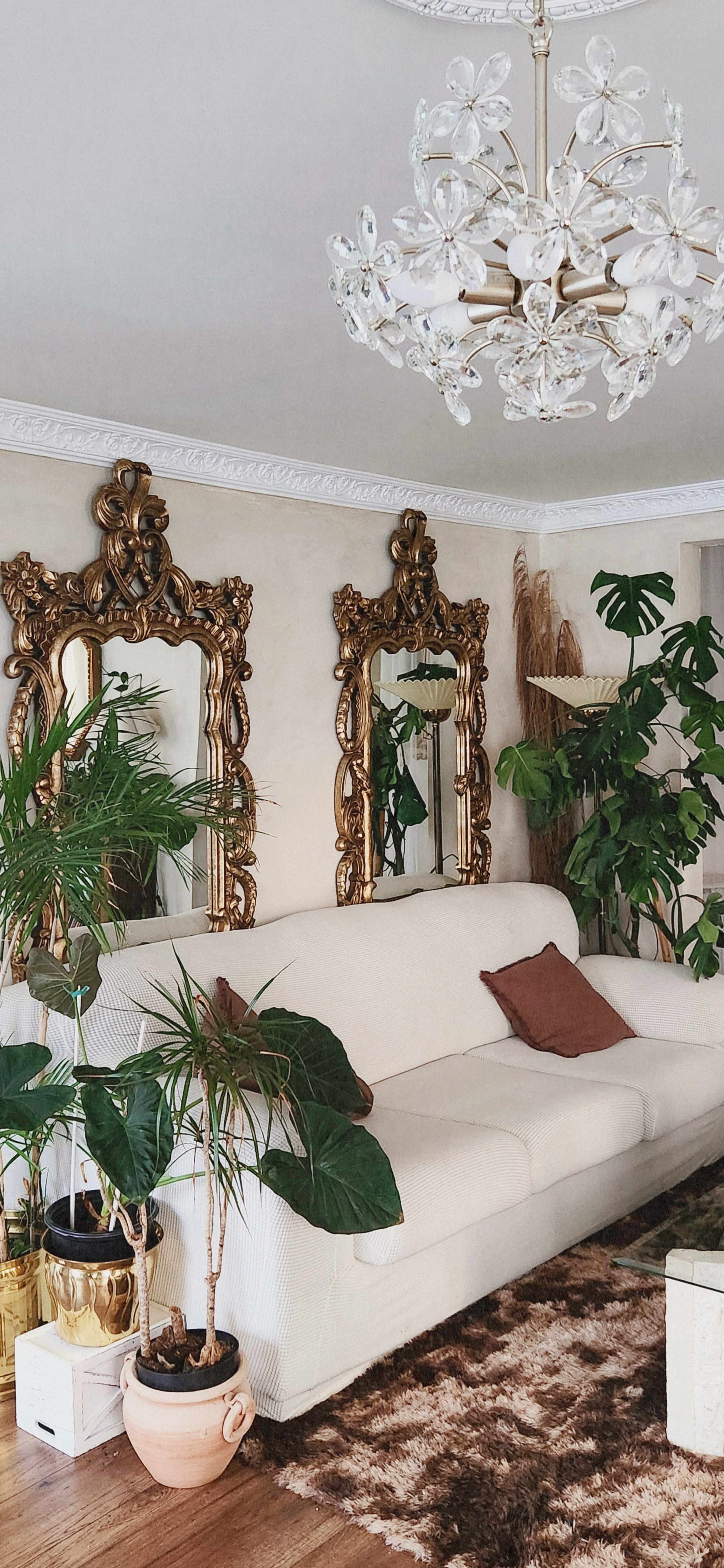 The image shows a living room with a white sofa, two ornate gold-framed mirrors mounted on the wall, various indoor plants, and a decorative chandelier.