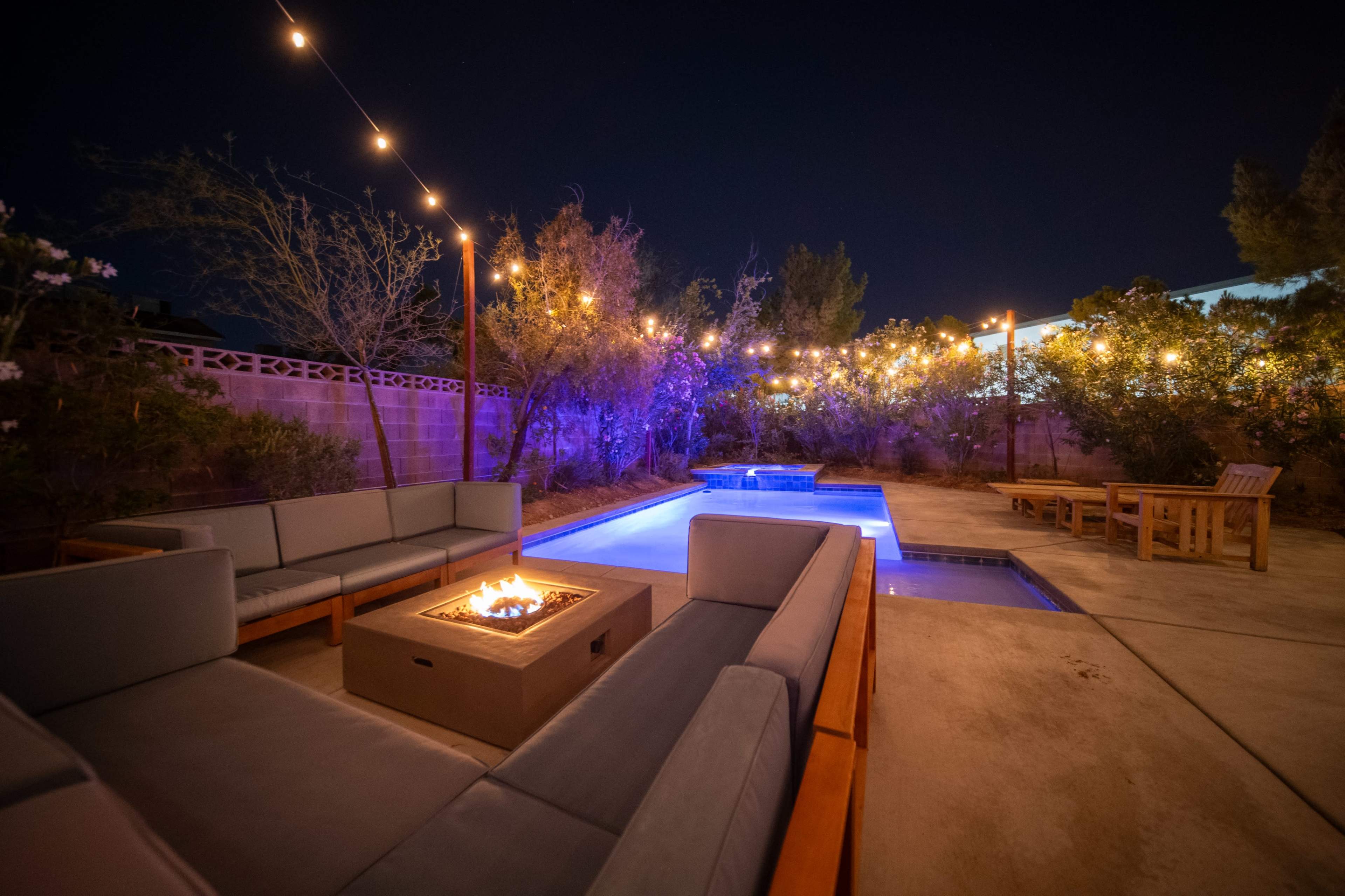 The image shows a backyard at night featuring a lit pool, a fire pit surrounded by modern seating, and string lights illuminating the surrounding plants.