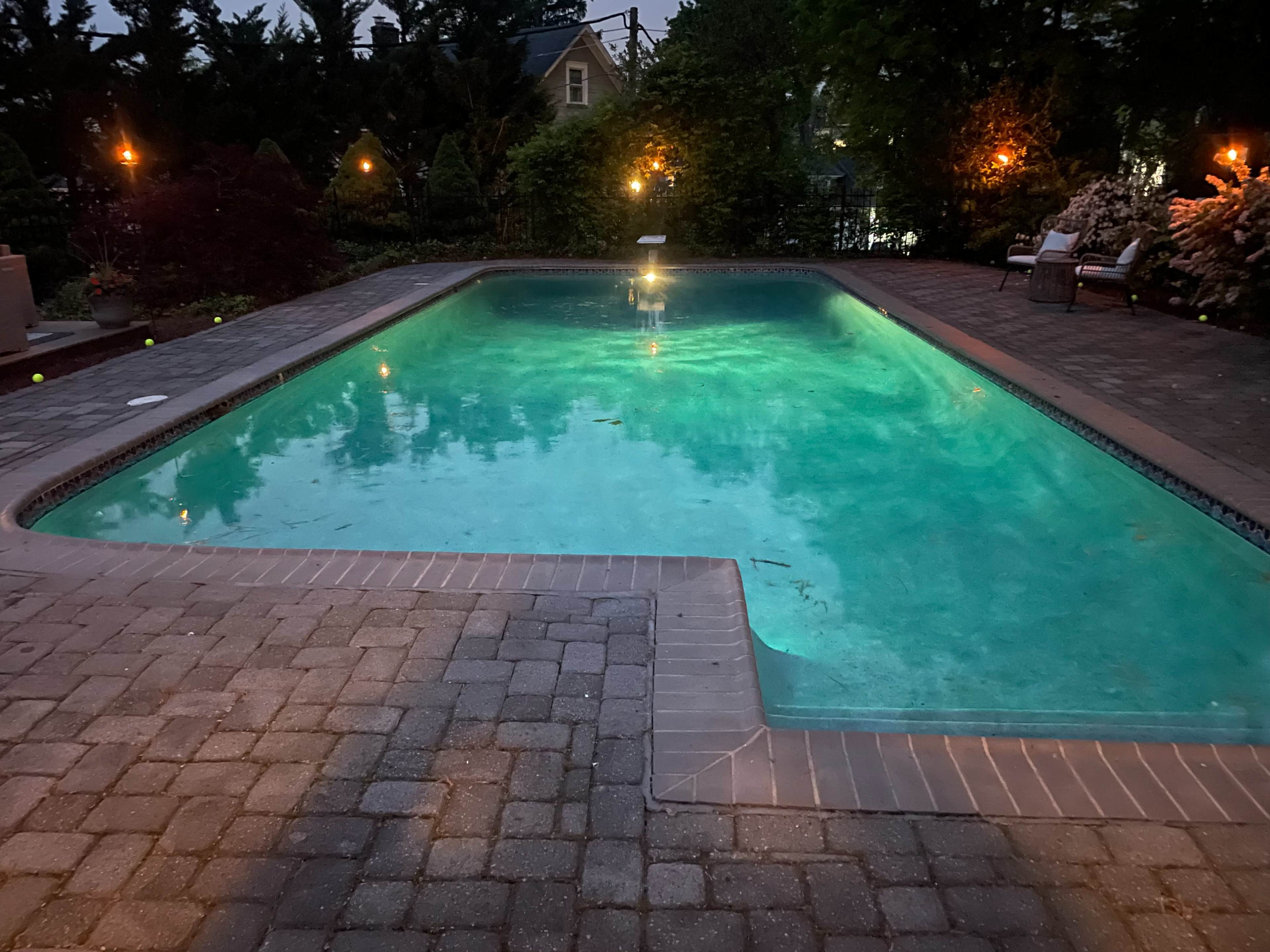 The image shows a lit swimming pool surrounded by a paved patio and landscaped greenery.