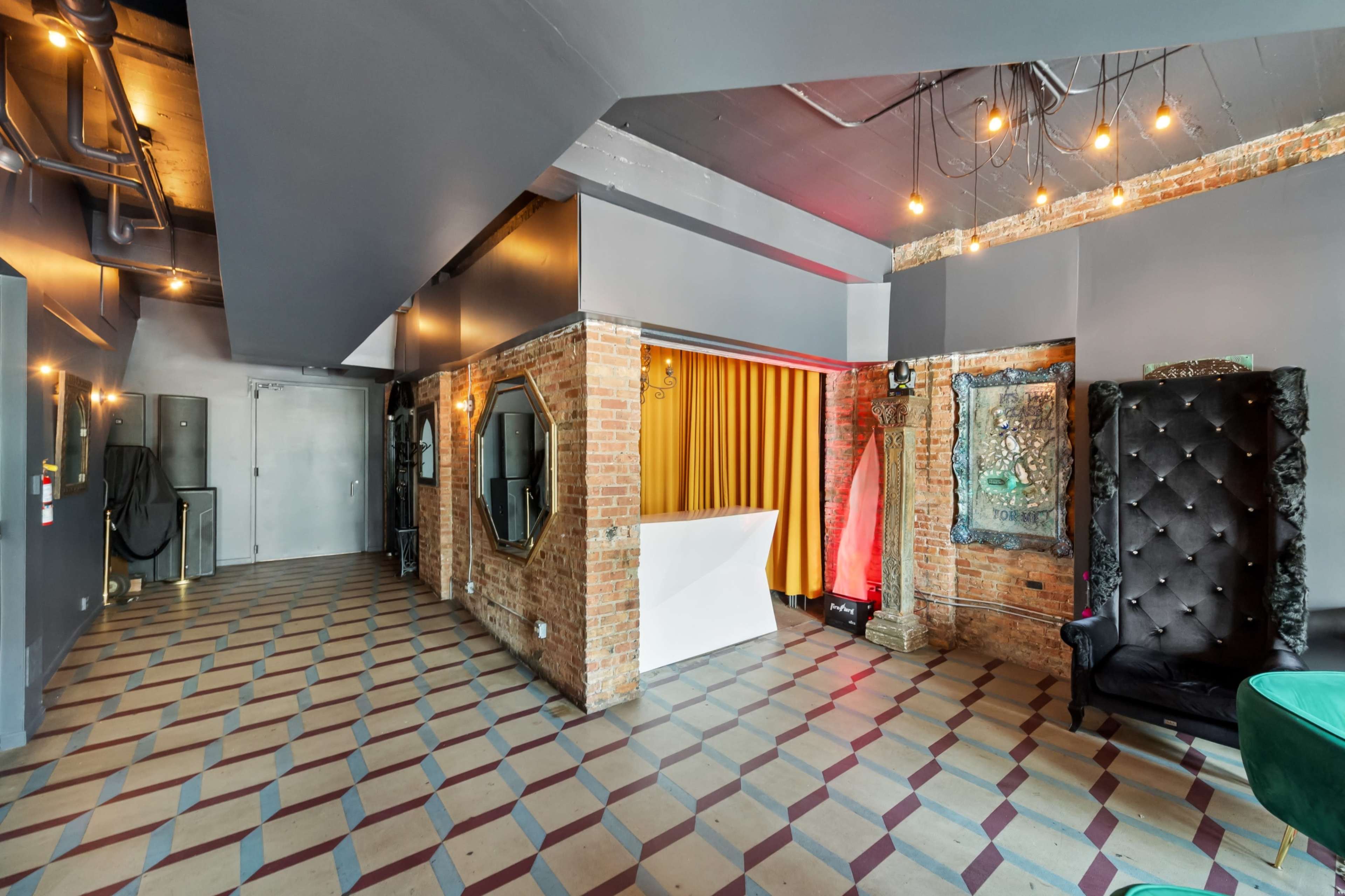 The image shows a contemporary interior space with a patterned tile floor, exposed brick walls, a bar area with a white counter, and stylish black seating against a backdrop of yellow drapery.