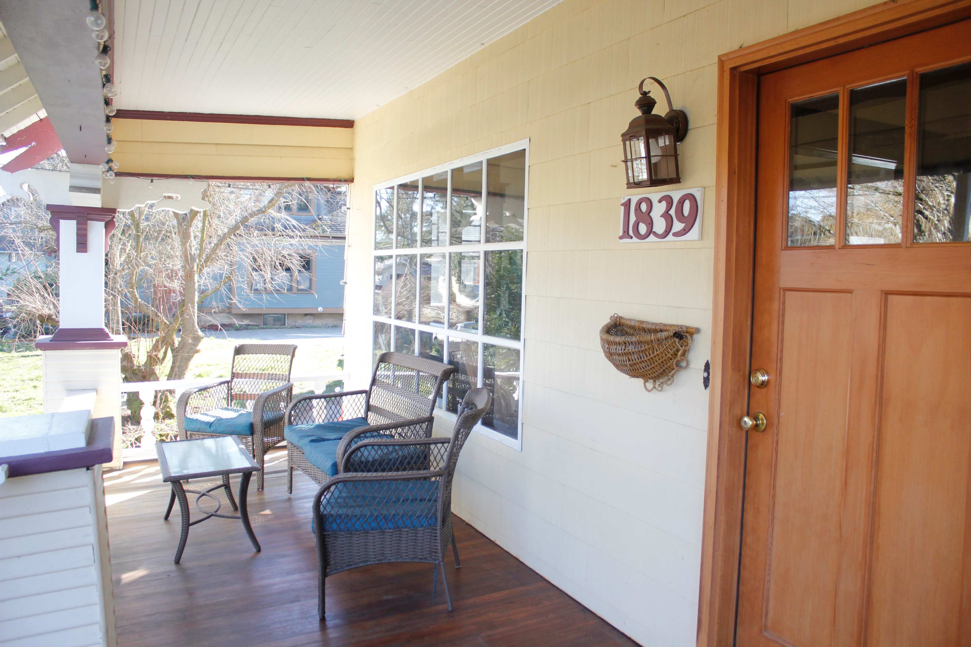 A porch with a wooden floor, a front door, a house number sign reading "1839," and seating arrangements including a table and chairs.