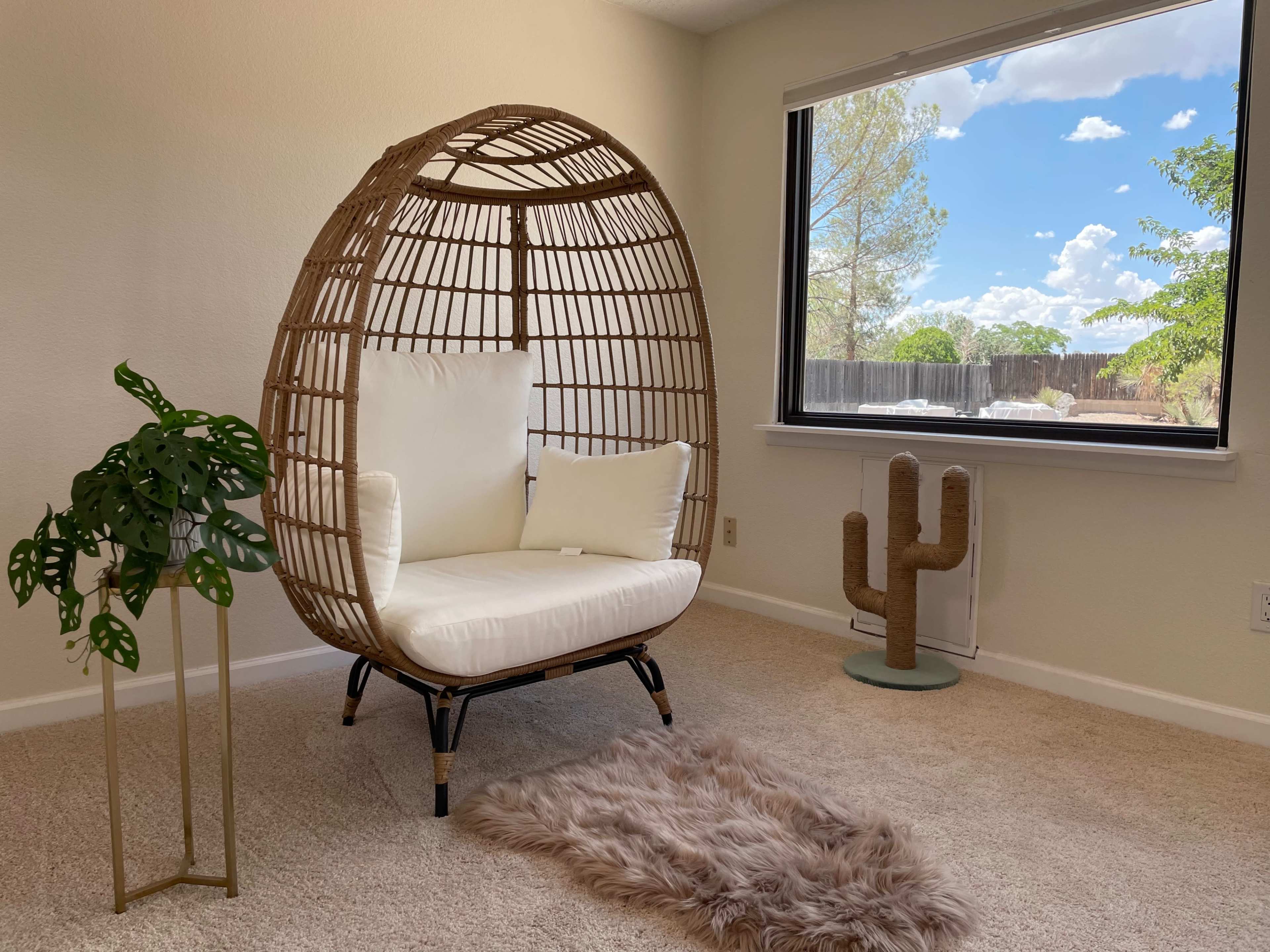 A wicker hanging chair with white cushions sits beside a potted plant and a decorative cactus in a well-lit room.