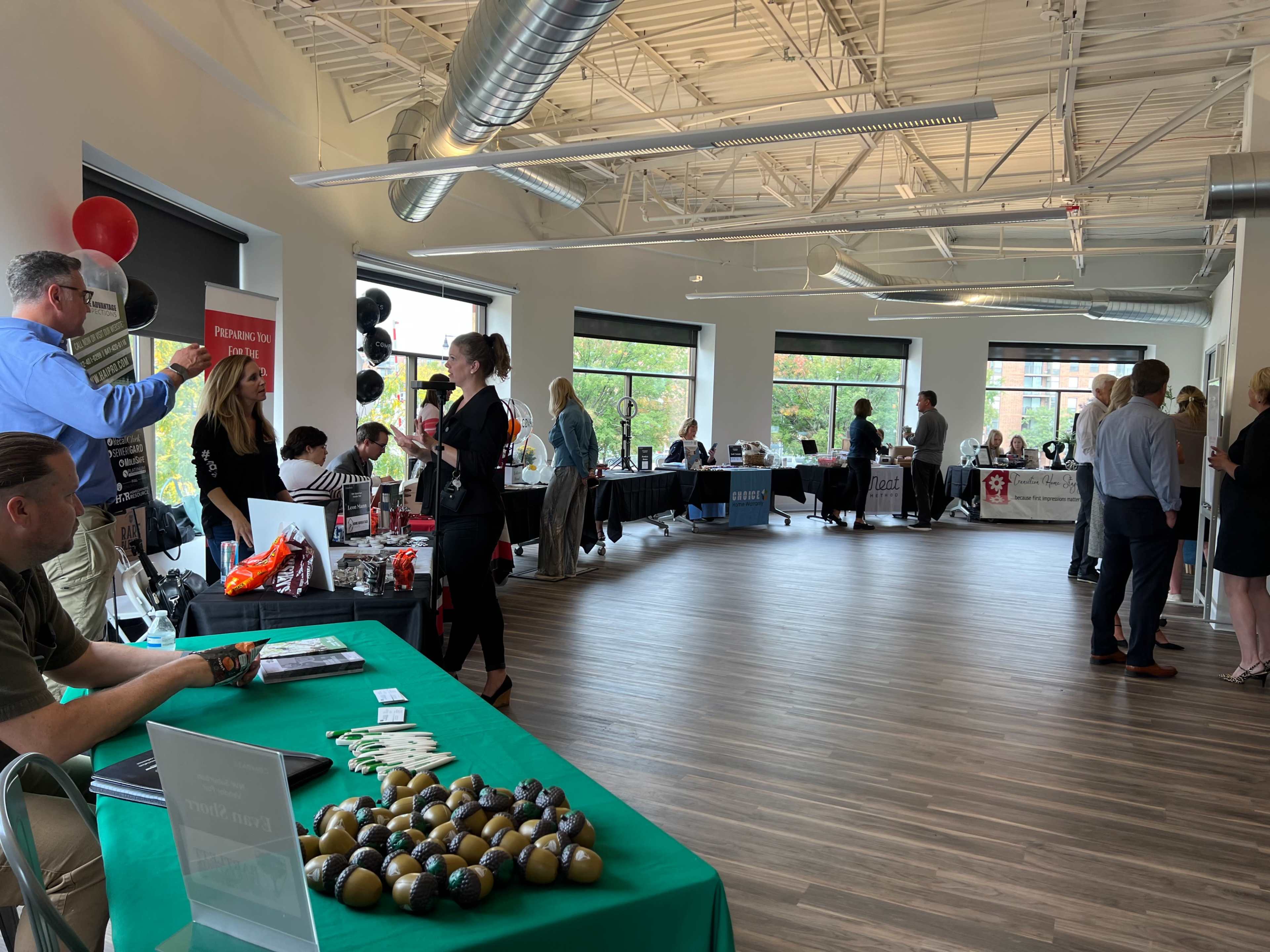 A spacious indoor event space is filled with various booths and attendees engaging in conversations, with tables displaying promotional materials and snacks.