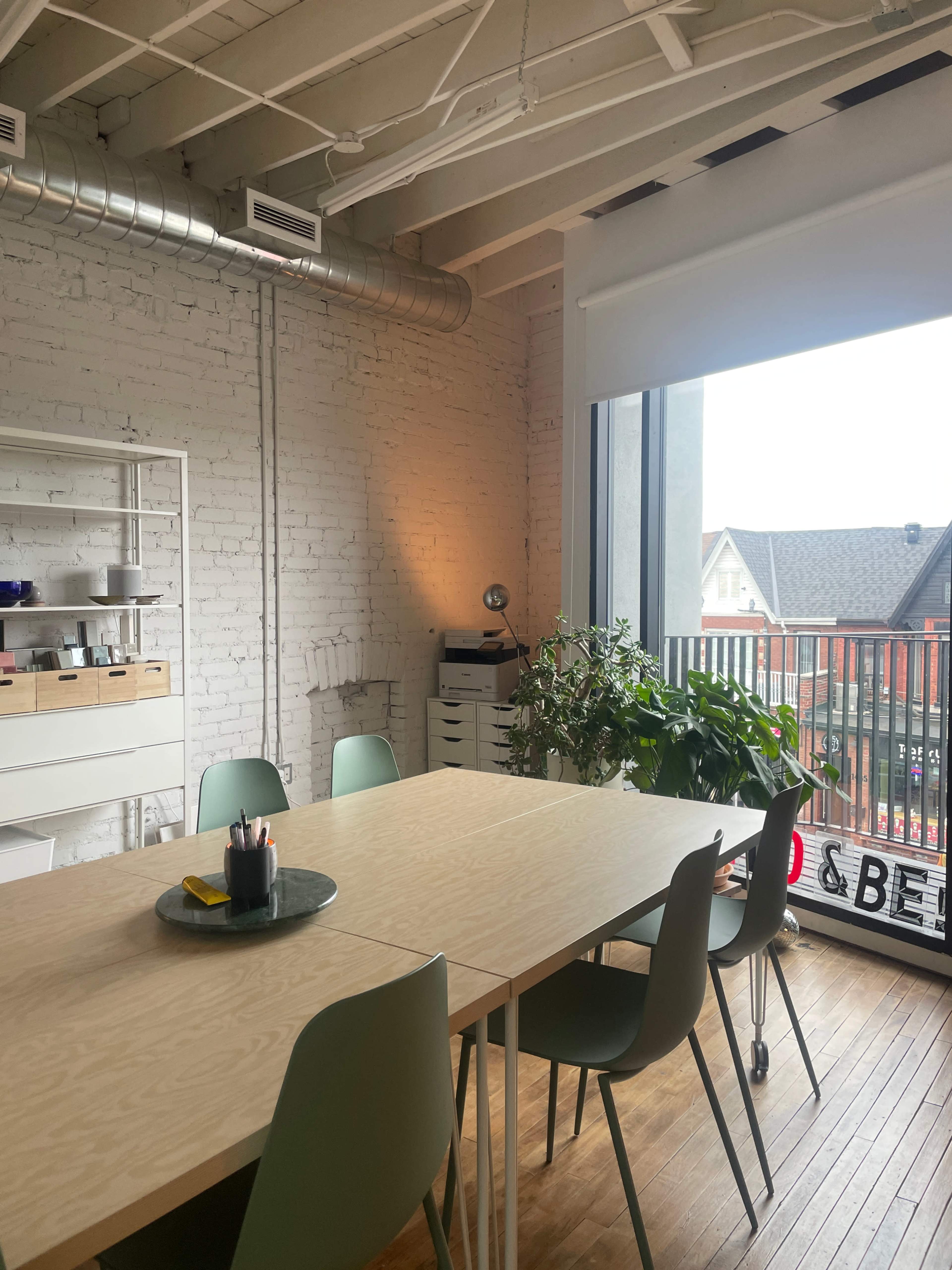 The image shows a modern meeting room with a large wooden table surrounded by green chairs, a plant, and large windows allowing natural light to enter the space.