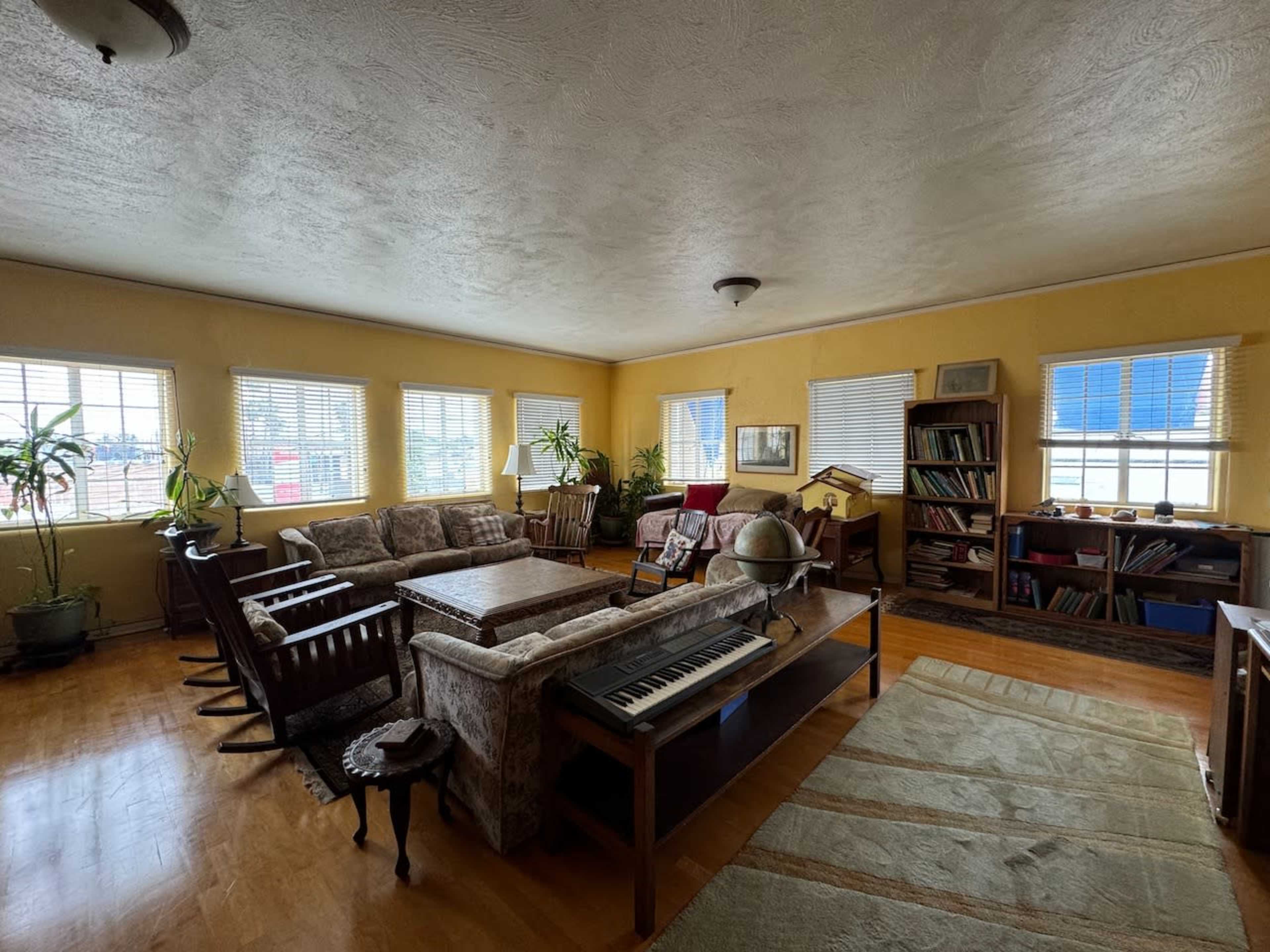 The image shows a spacious, warmly-lit room filled with various seating arrangements, plants, and bookshelves.