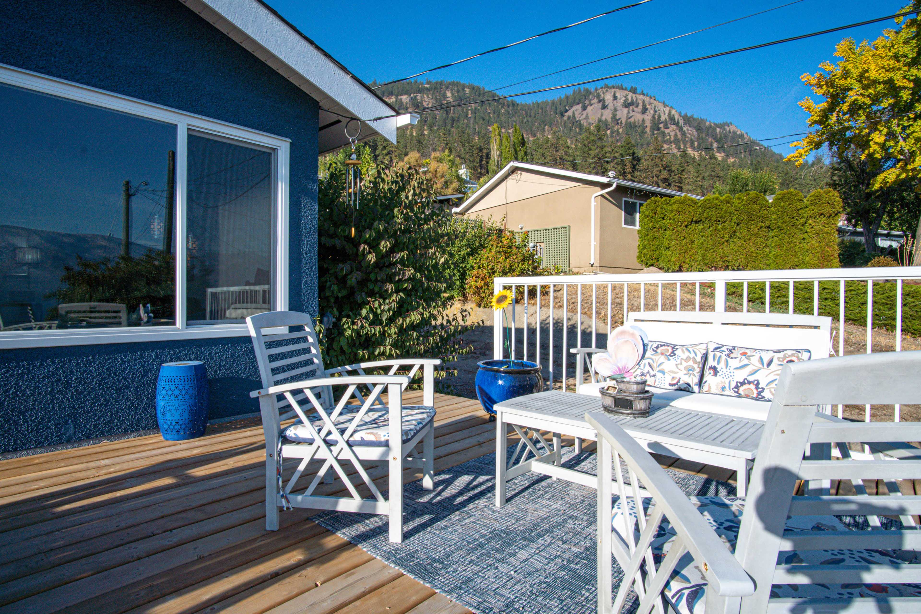 A deck features white outdoor furniture and colorful planters, surrounded by mountains and houses under a blue sky.