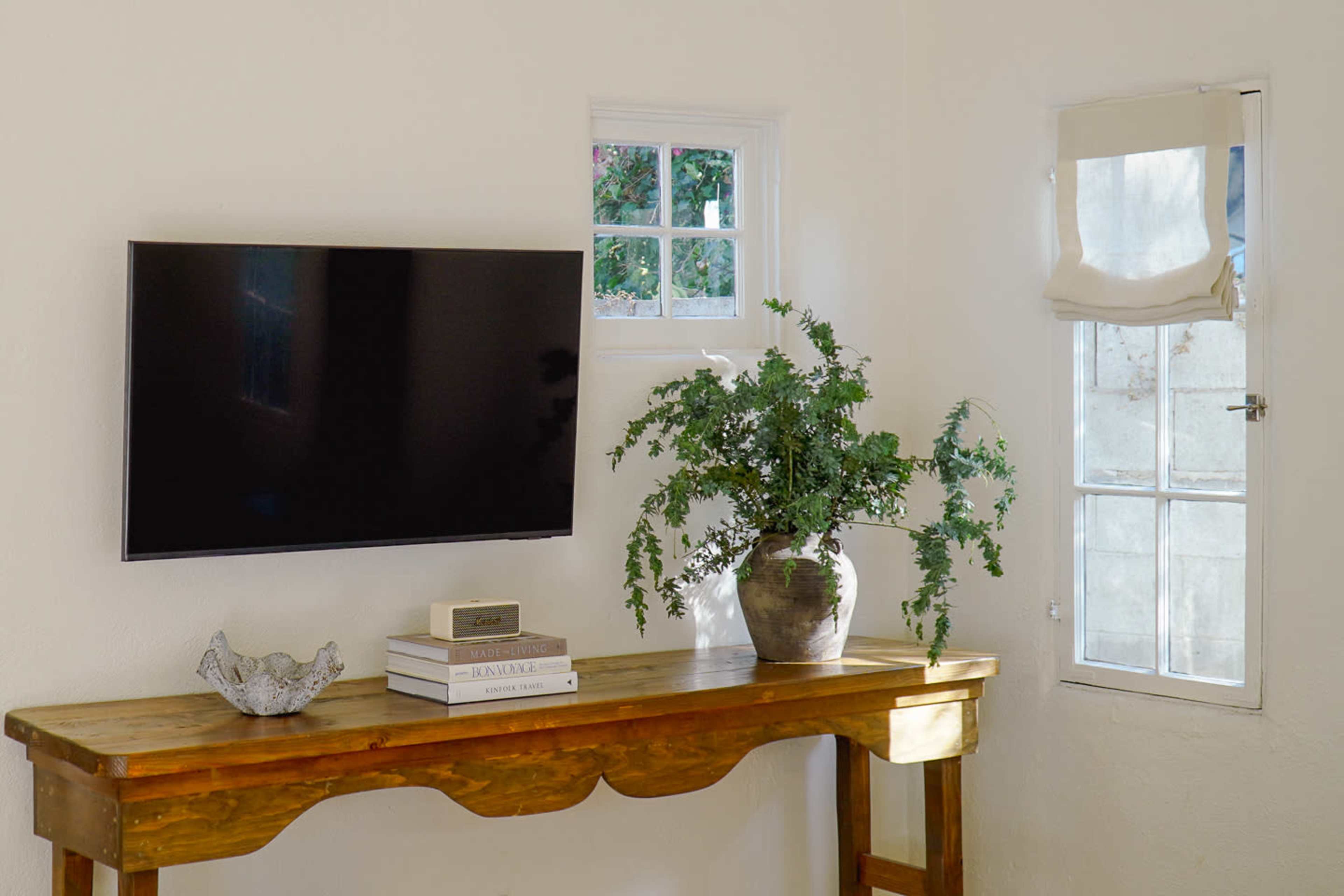 A wall-mounted television is displayed above a wooden console table adorned with stacked books and a potted plant, beside a window with a light fabric cover.