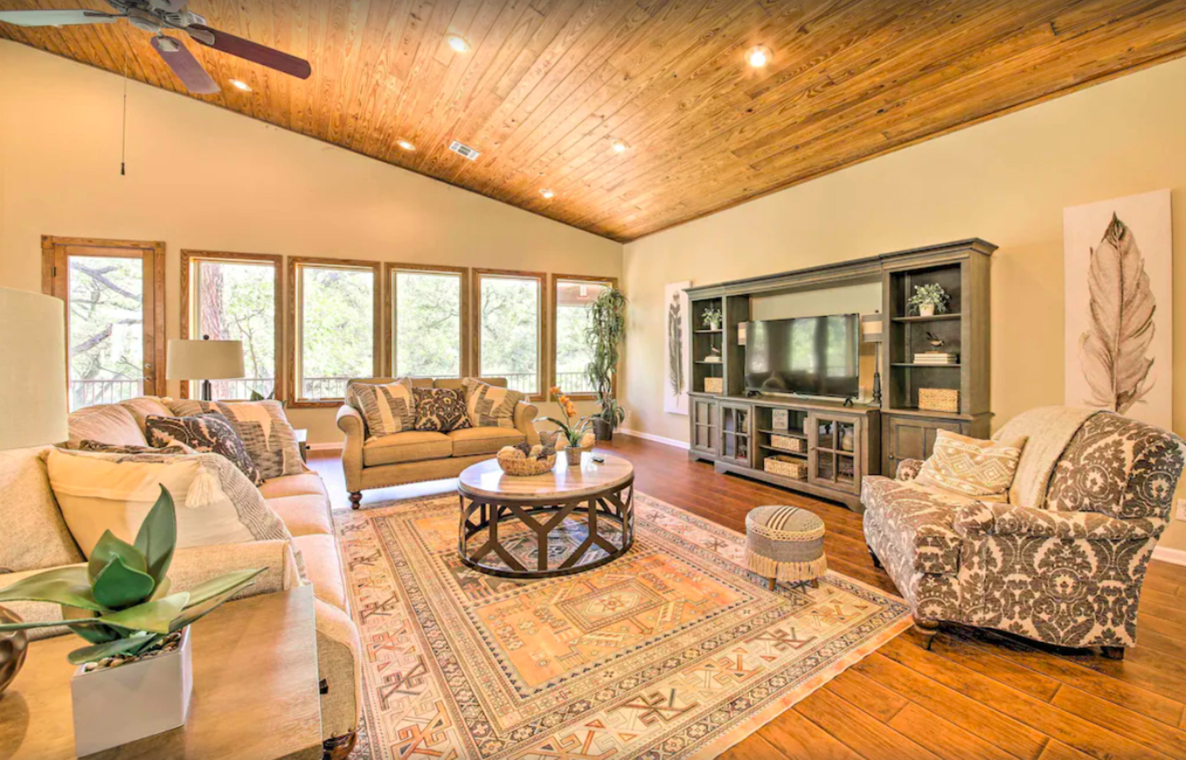 The living room features a large area rug, a circular coffee table, and a media console against a wall of windows that offer a view of the outdoors.