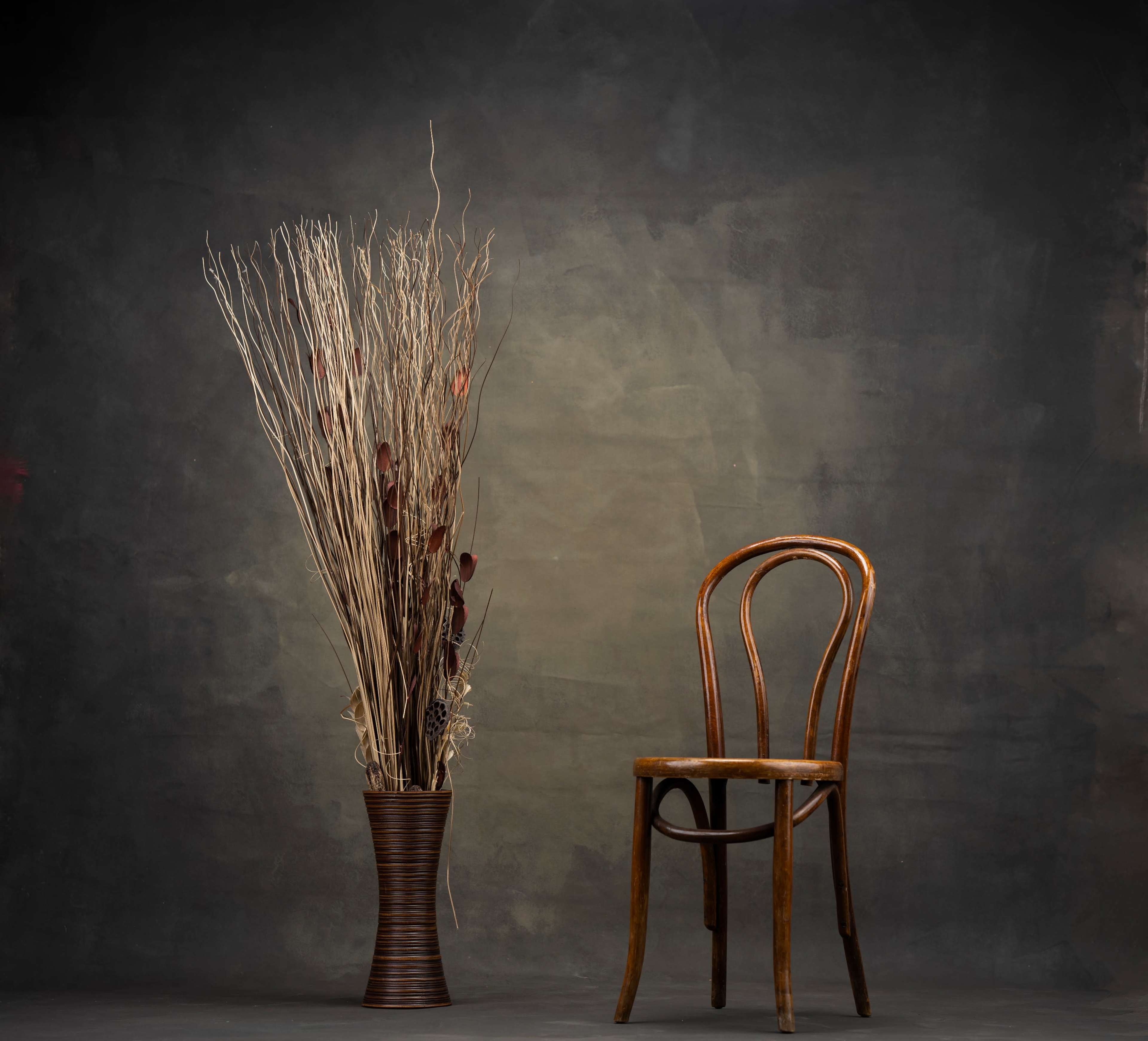 A wooden chair stands beside a tall vase filled with decorative branches against a textured backdrop.