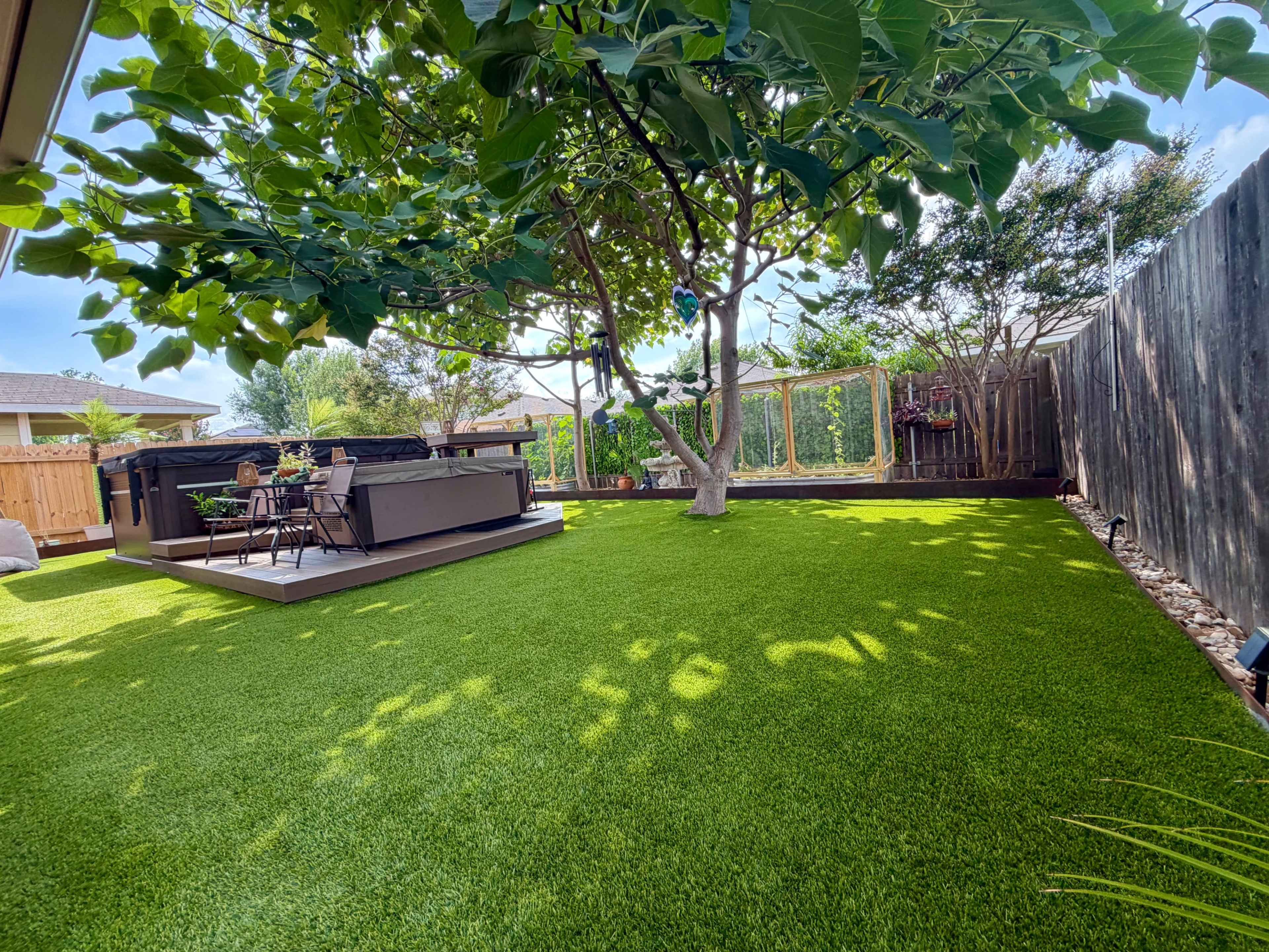 The image shows a spacious backyard with artificial grass, a deck with patio furniture, and trees providing shade.